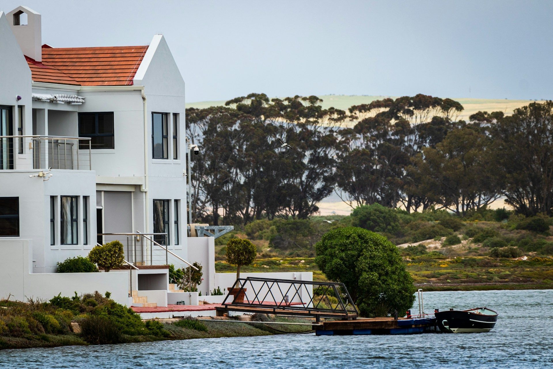 White houses on a waterfront with a pier, trees and a dune on the background, blue water.