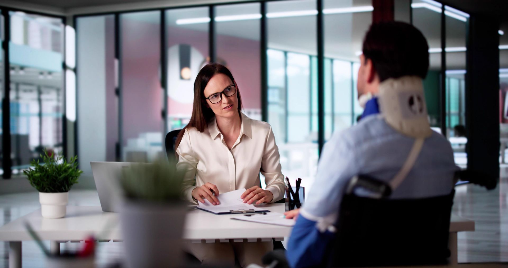 Woman in glasses talking to person in wheelchair wearing neck brace at an office desk.