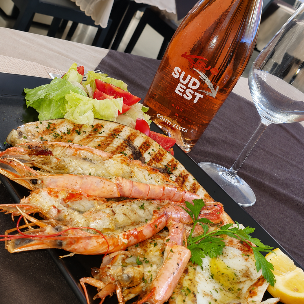 Piatto di frutti di mare grigliati con gamberi, limone, insalata e una bottiglia di rosé su un tavolo.