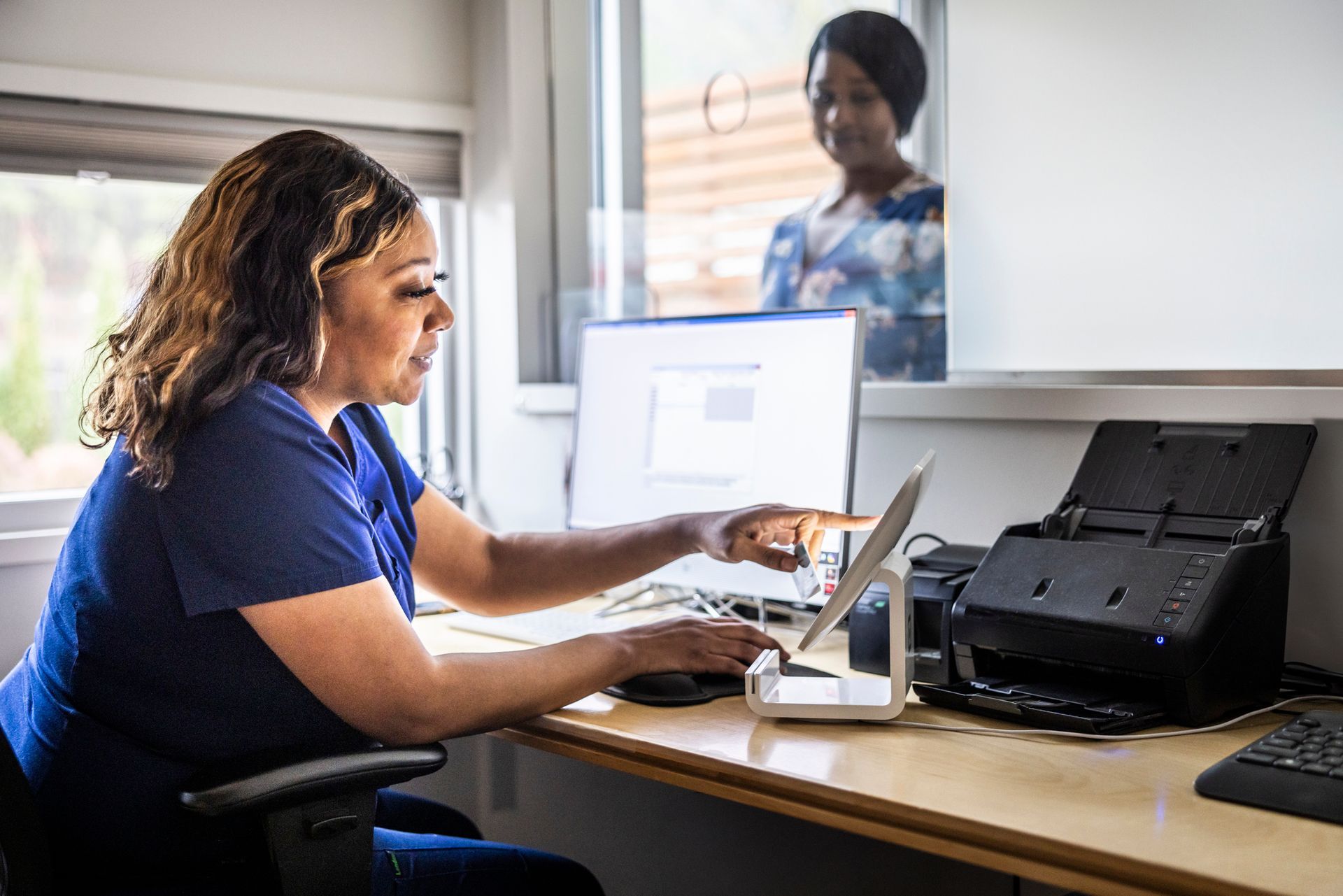 Woman in blue scrubs points at tablet, sitting at desk. Another woman looks on from the window.