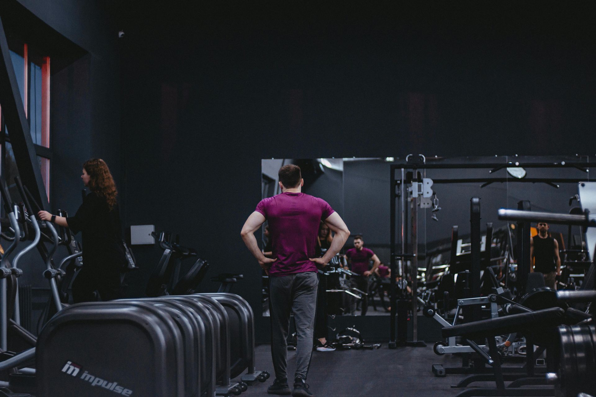 People exercising in a dark gym, with a man in a magenta shirt standing near workout equipment.