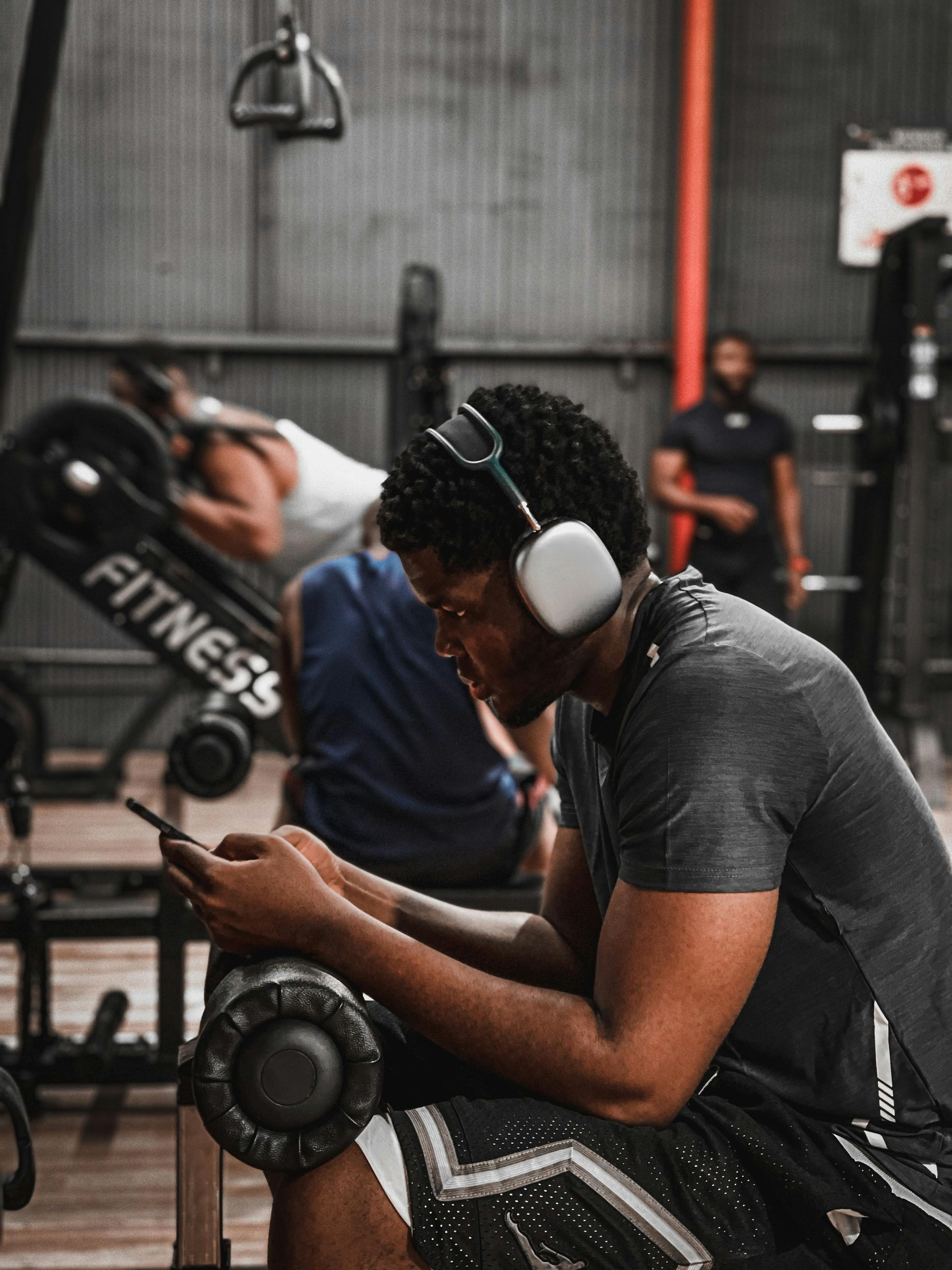 Person resting at a gym machine, wearing headphones, with weights and another exerciser in the background.