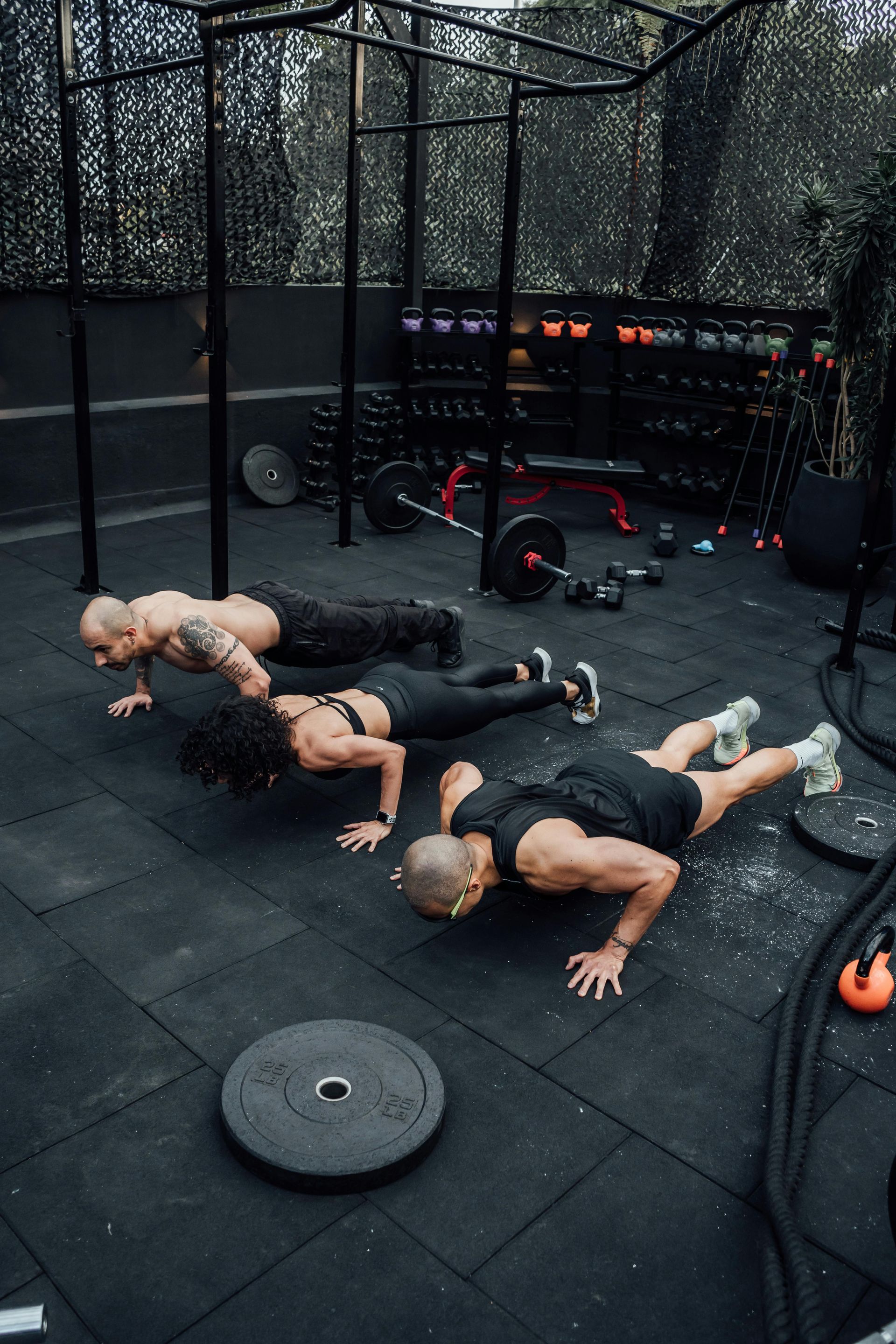 Four people doing push-ups on a black outdoor gym mat with kettlebells and chain-link fencing nearby
