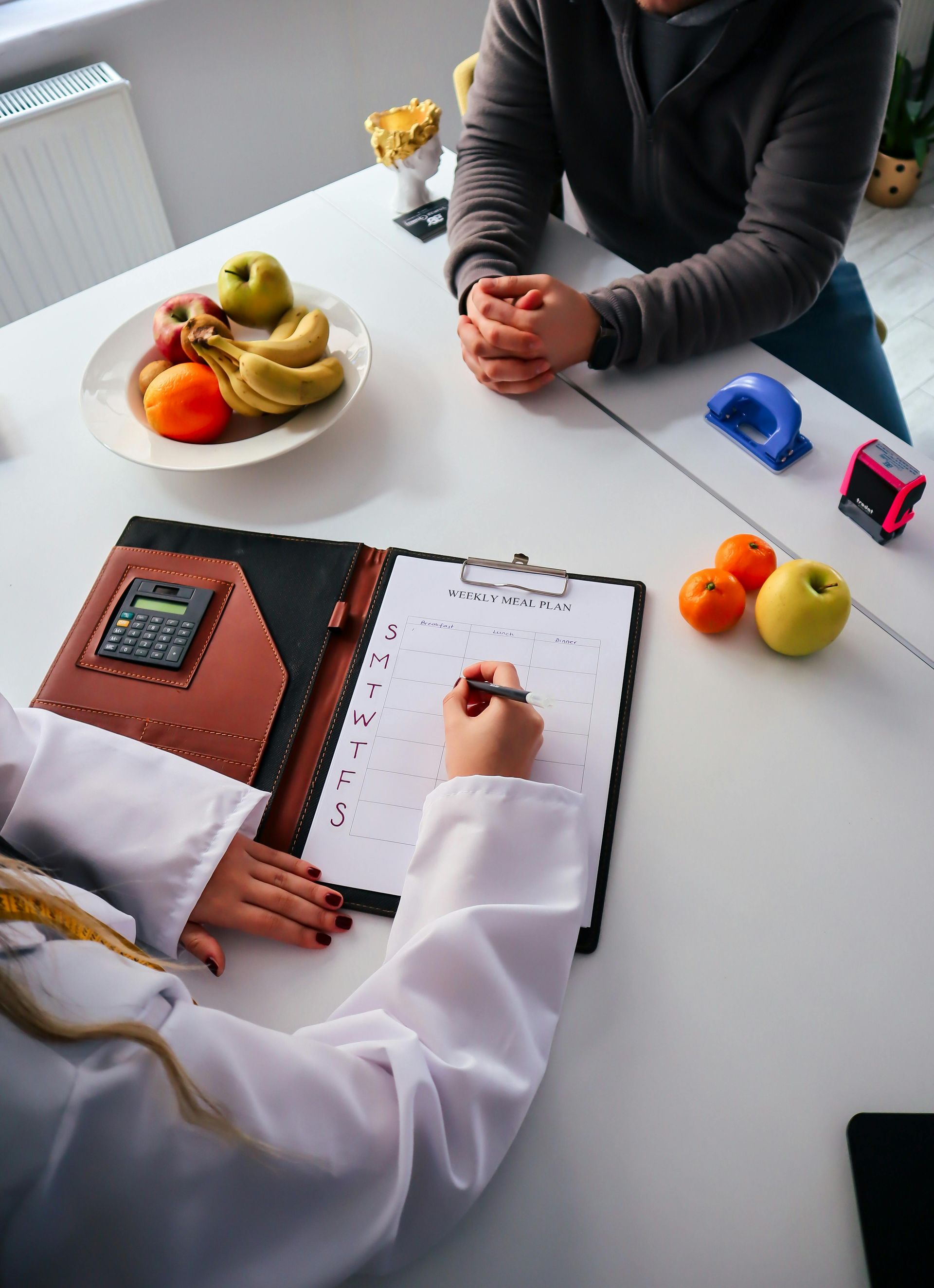Two people review a notebook at a white table with fruit bowls, a blue tape dispenser, and a pen.