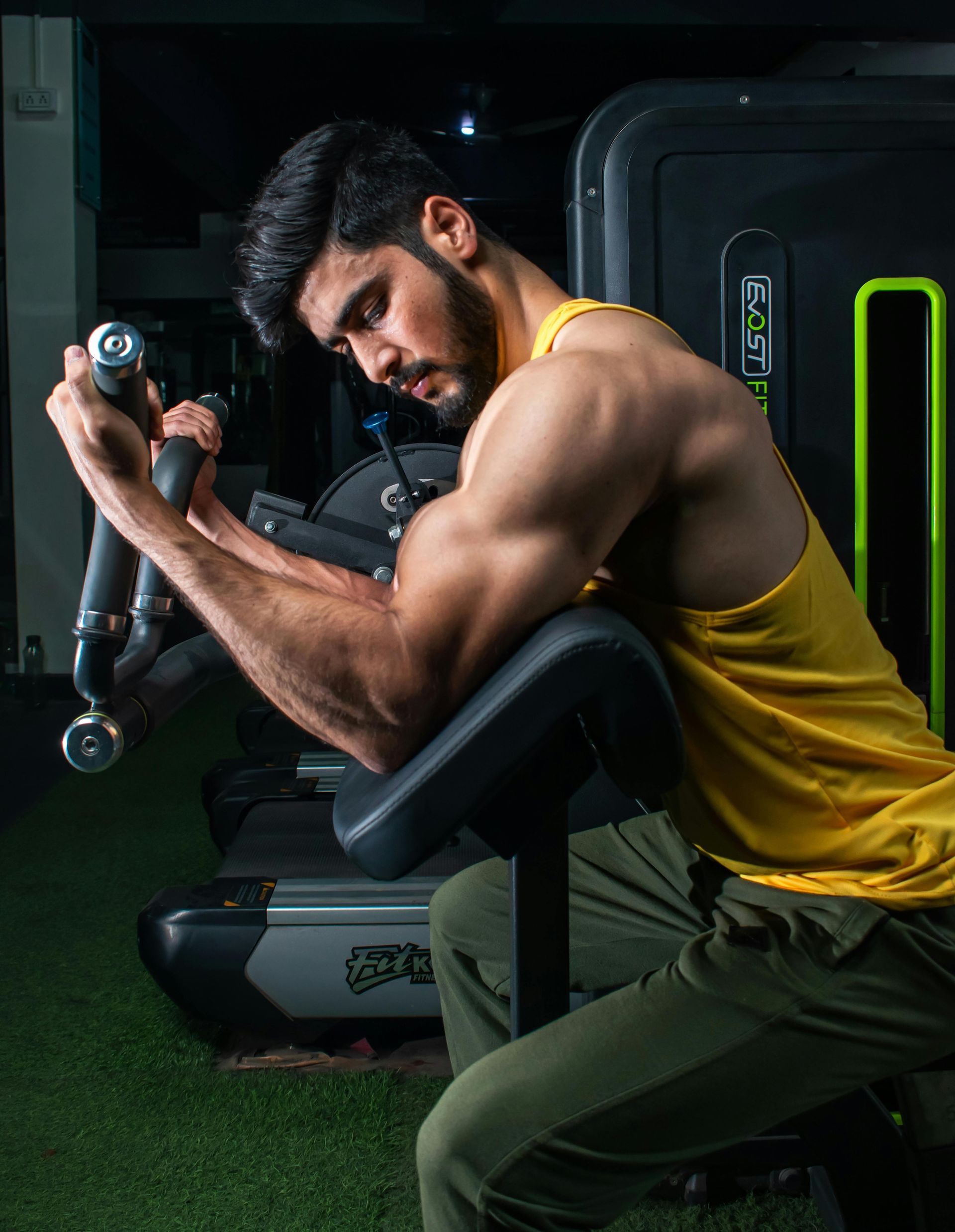 Man flexing biceps on a gym machine, wearing a yellow tank top and green pants.