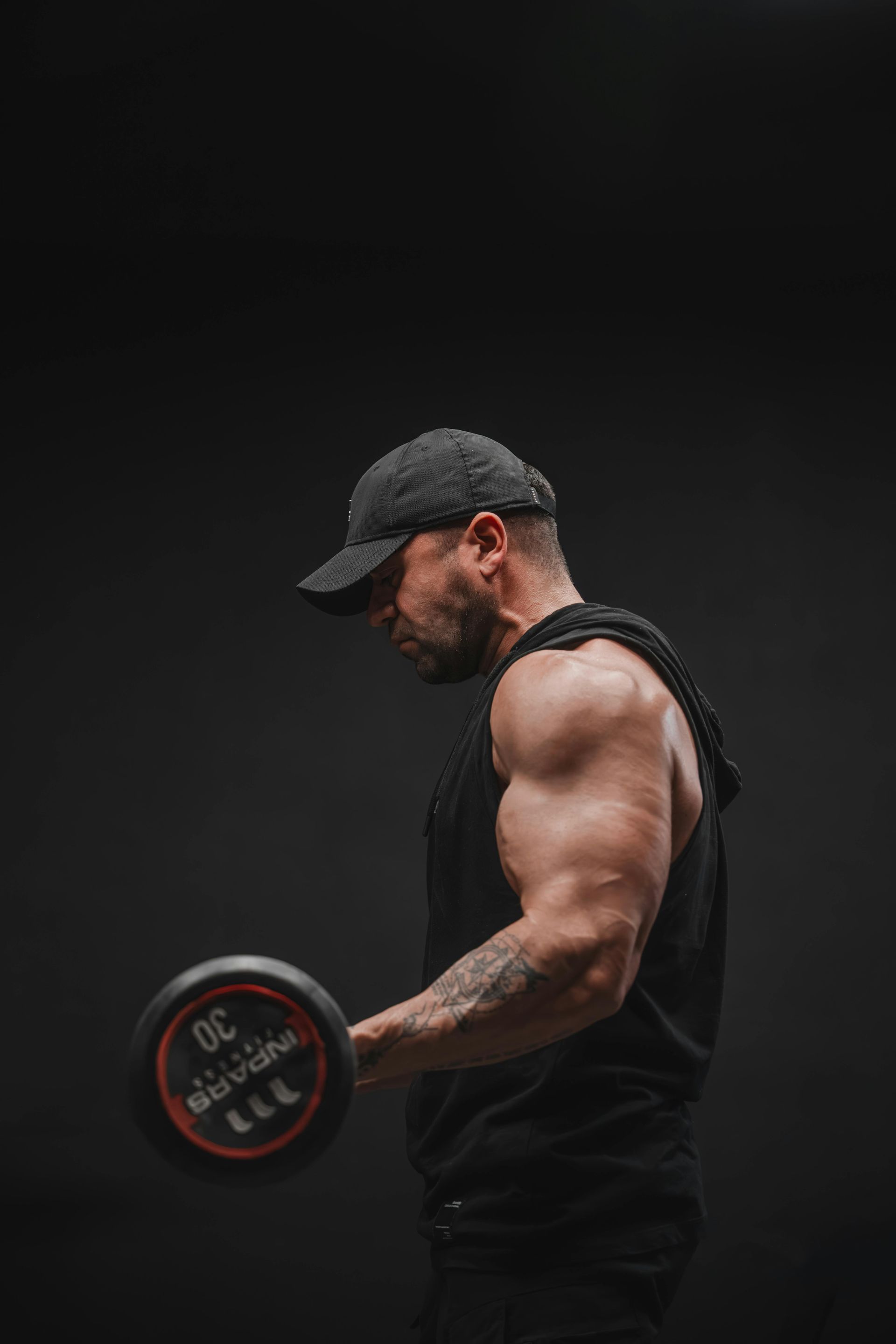 Muscular man in black tank top and cap doing a dumbbell curl against a dark background