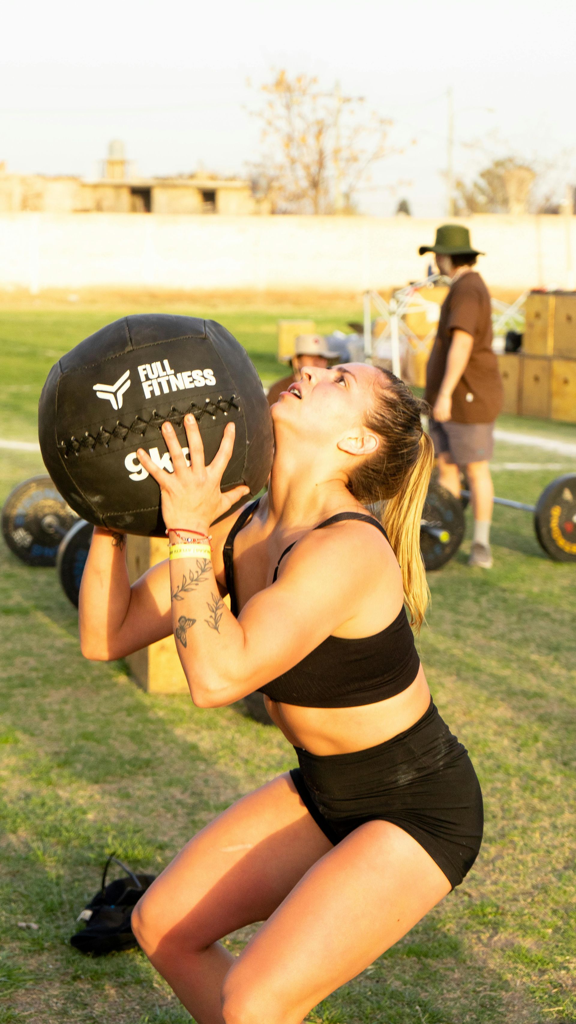 Woman in black athletic wear lifting a medicine ball outdoors on a grassy field at sunset