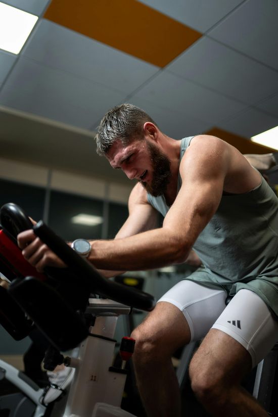Man pedaling a stationary bike in a gym, leaning forward and sweating under fluorescent lights