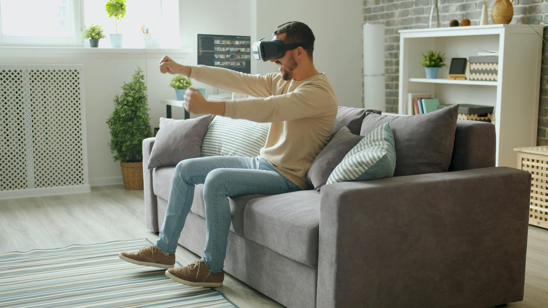 Person wearing a VR headset and holding controllers on a gray sofa in a bright living room.