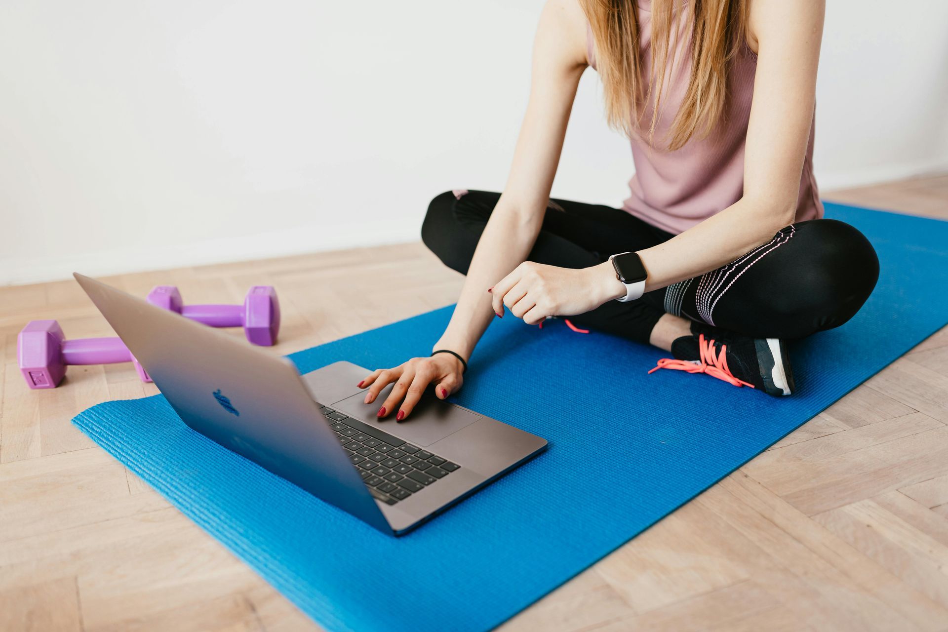 Person sitting on a blue yoga mat, using a laptop beside purple dumbbells in a bright room