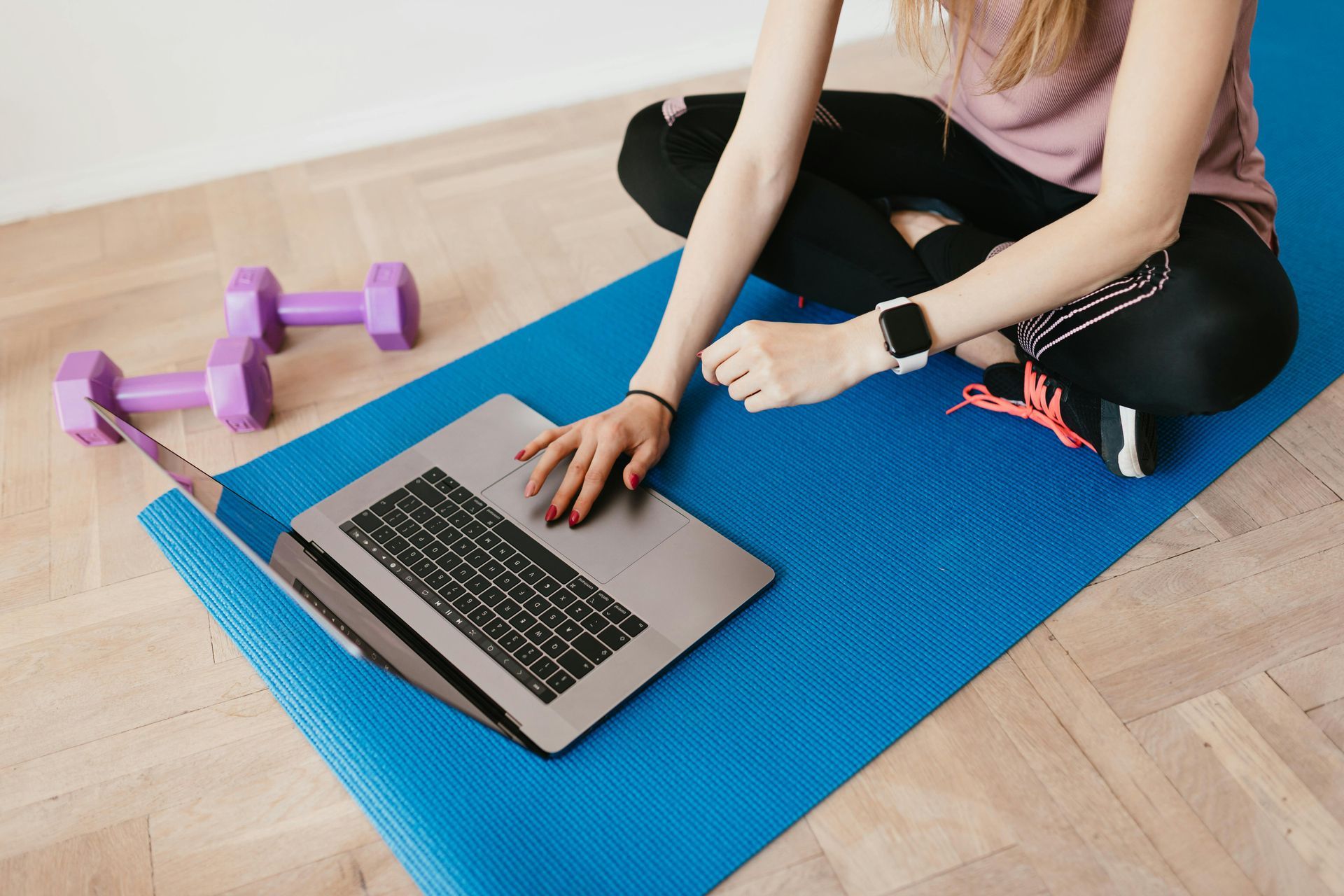 Person sitting on a blue yoga mat using a laptop beside purple dumbbells