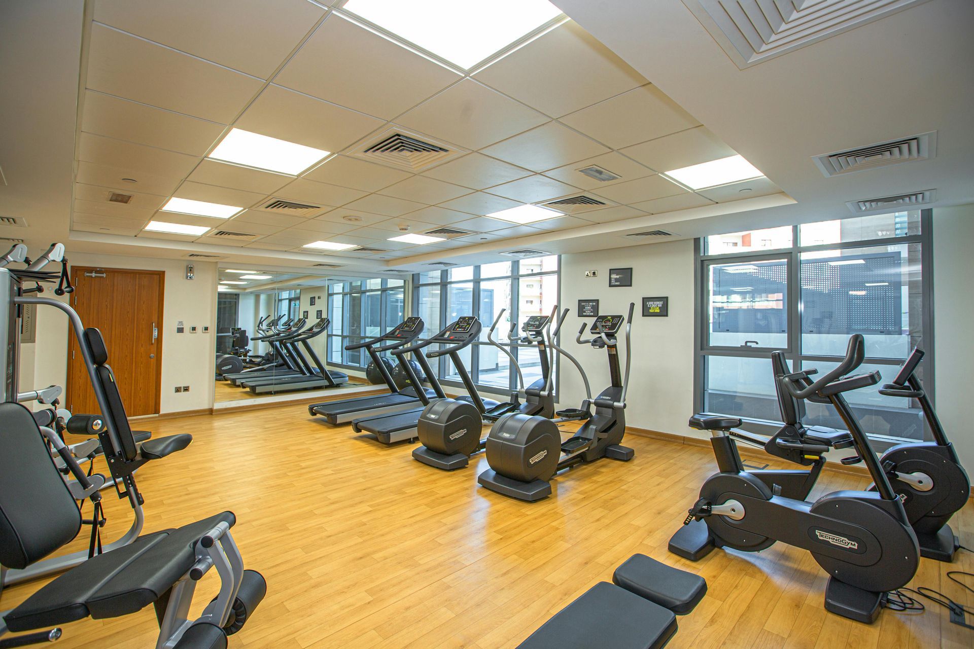 Bright gym with cardio machines, mirrored walls, and wood floors under ceiling lights.