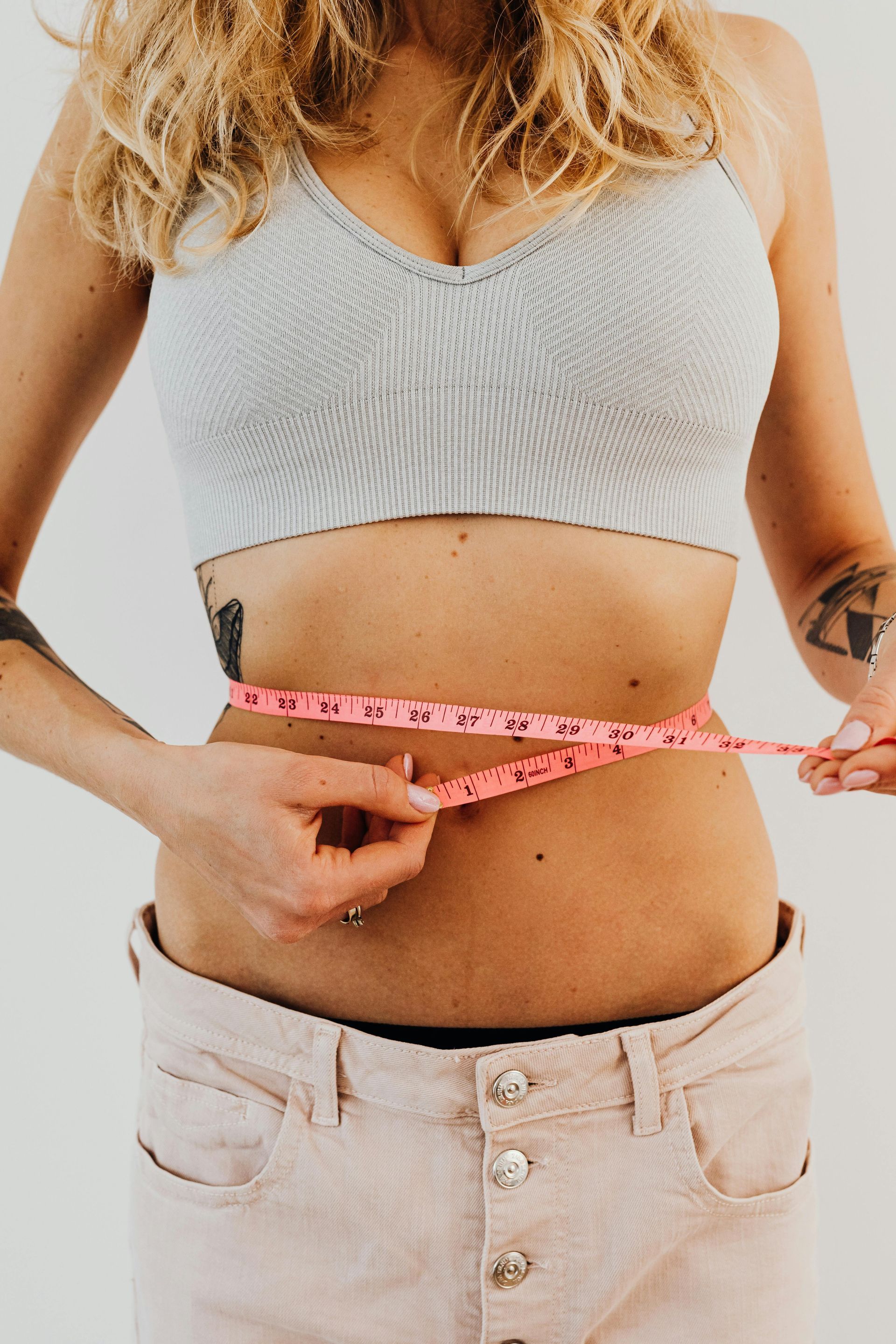 Person measuring waist with pink tape measure, wearing a gray crop top and light jeans.