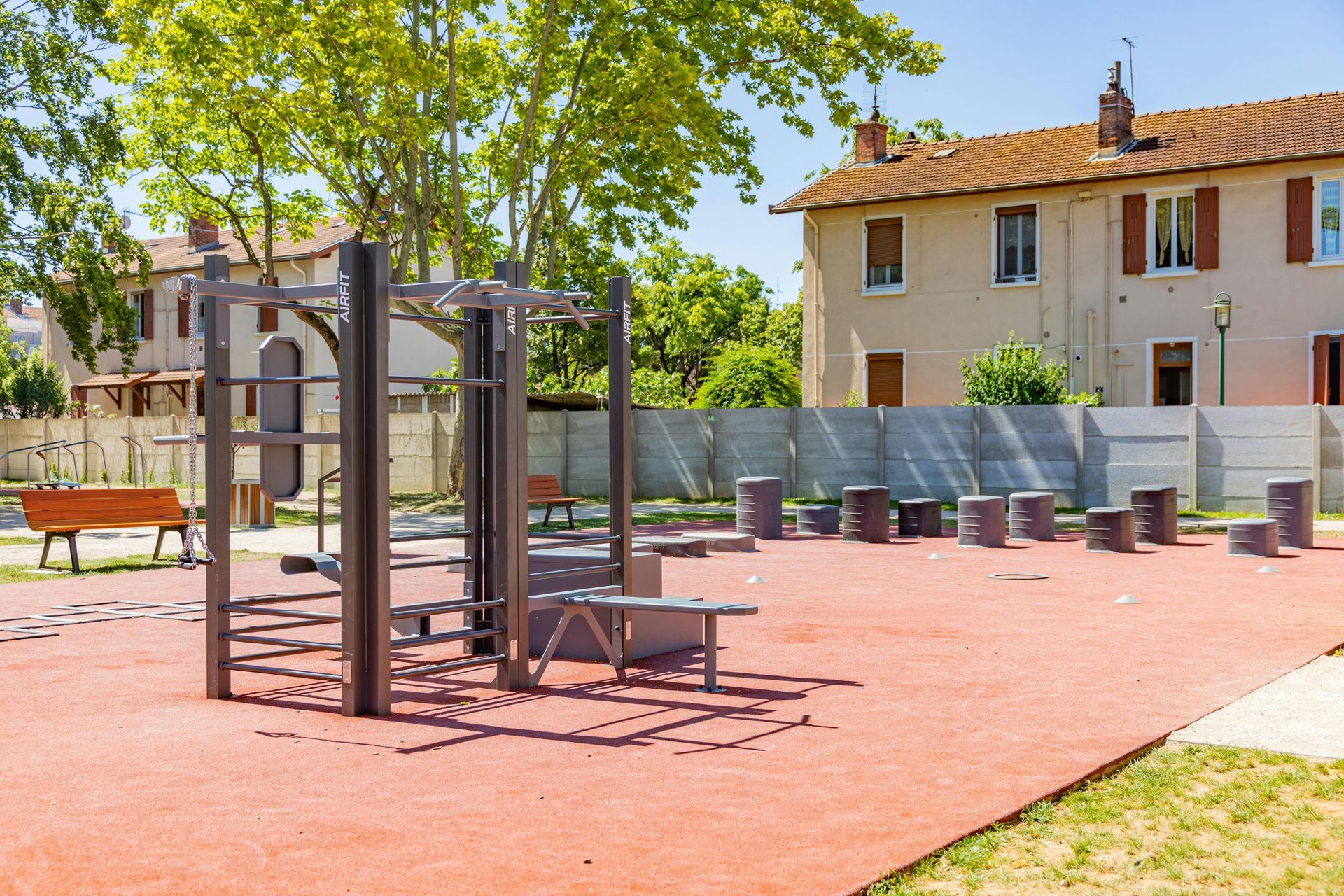 Outdoor playground with swings, benches, and a climbing frame on a red surface beside houses and trees