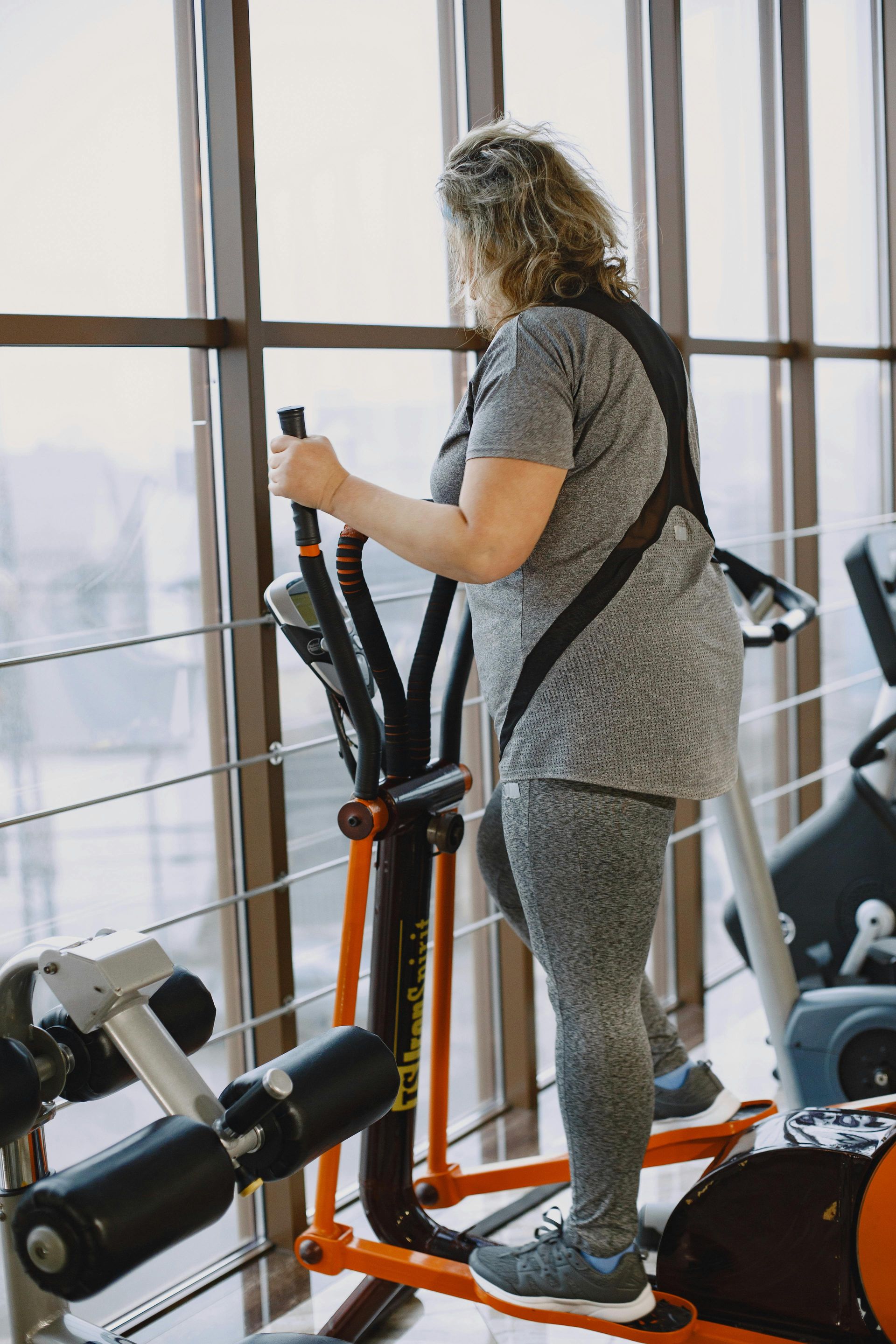 Person using an elliptical machine in a bright gym by large windows.
