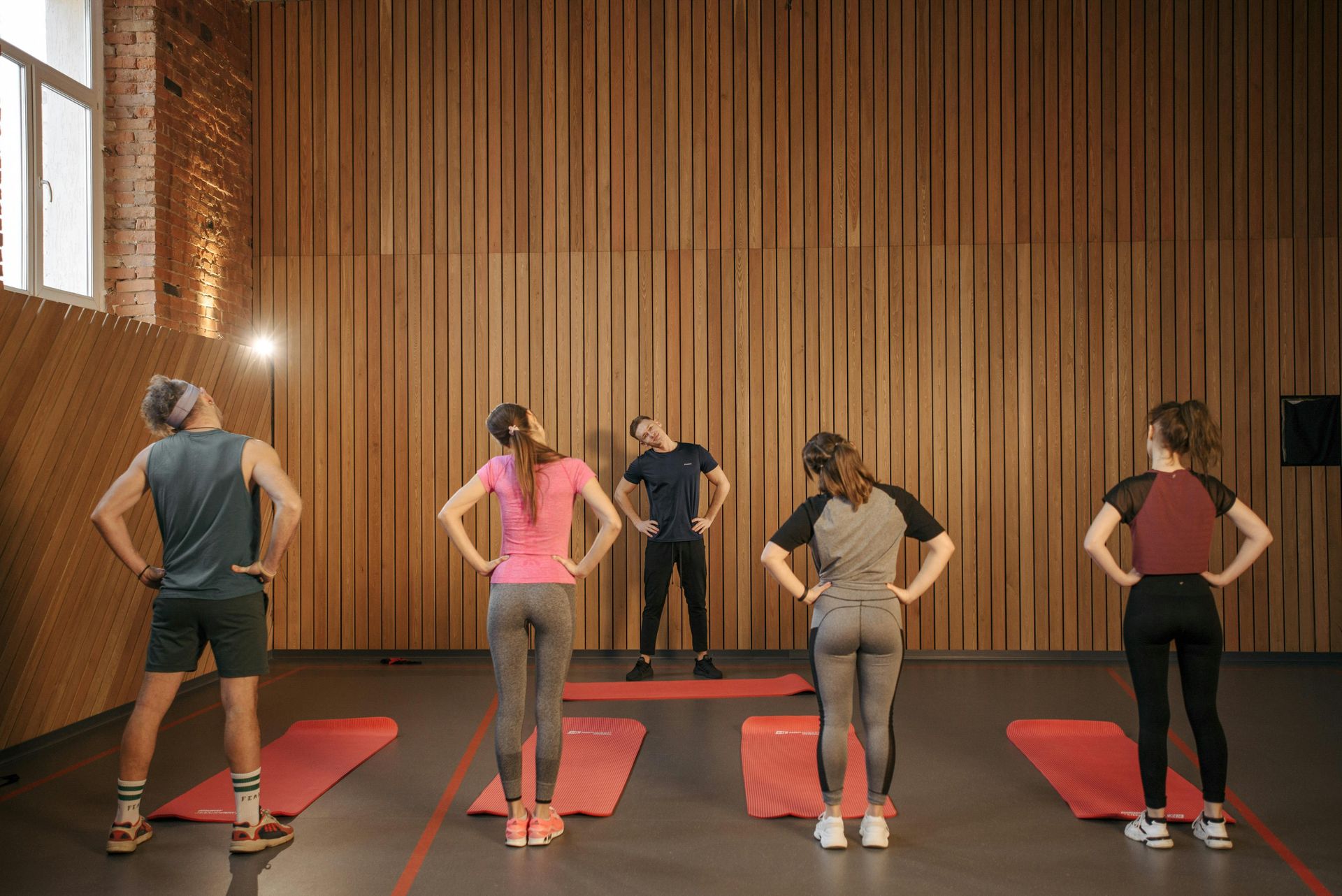 People standing on red exercise mats in a studio, facing a wooden wall during a workout class.