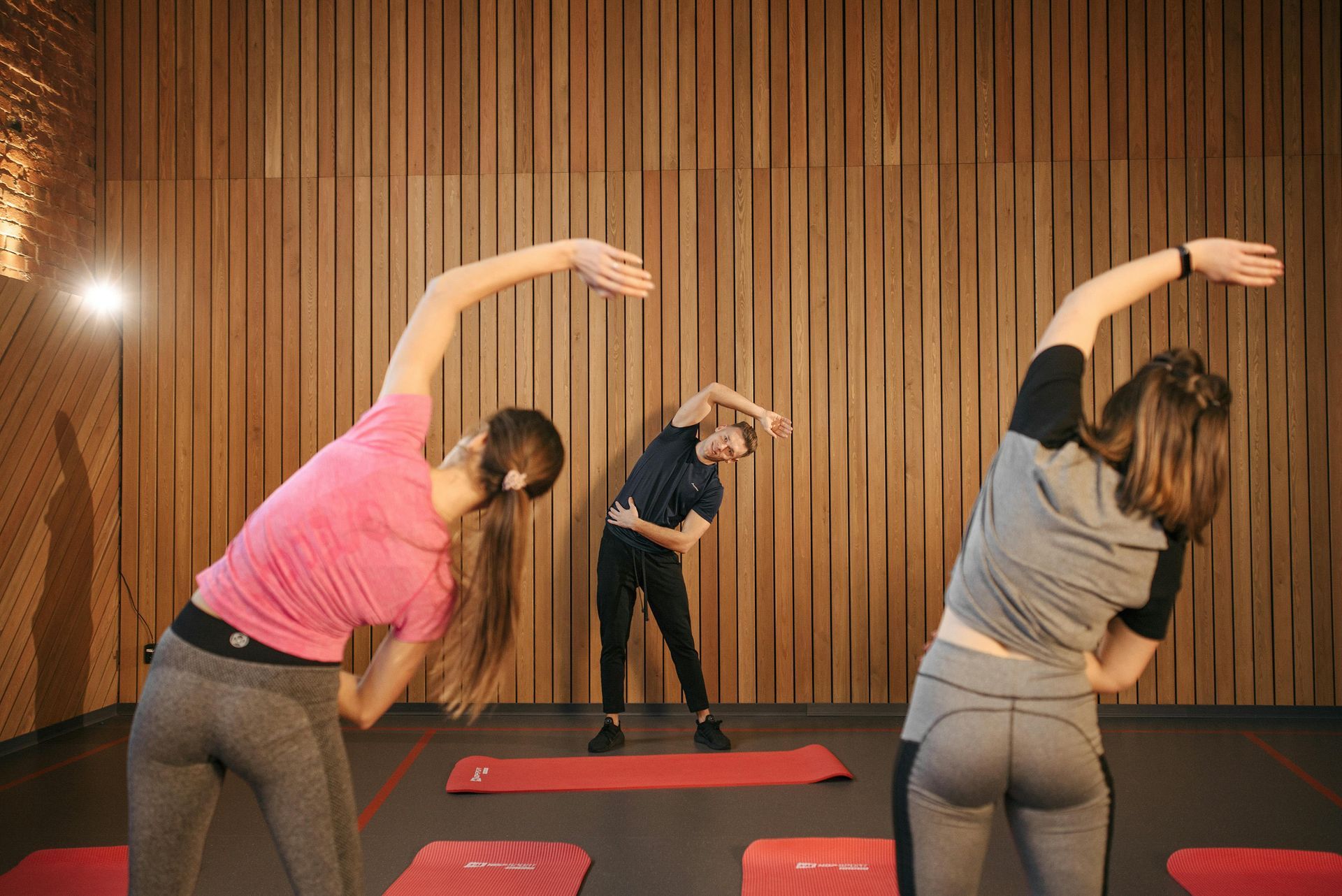 People stretching in a yoga class on red mats against a wooden wall.