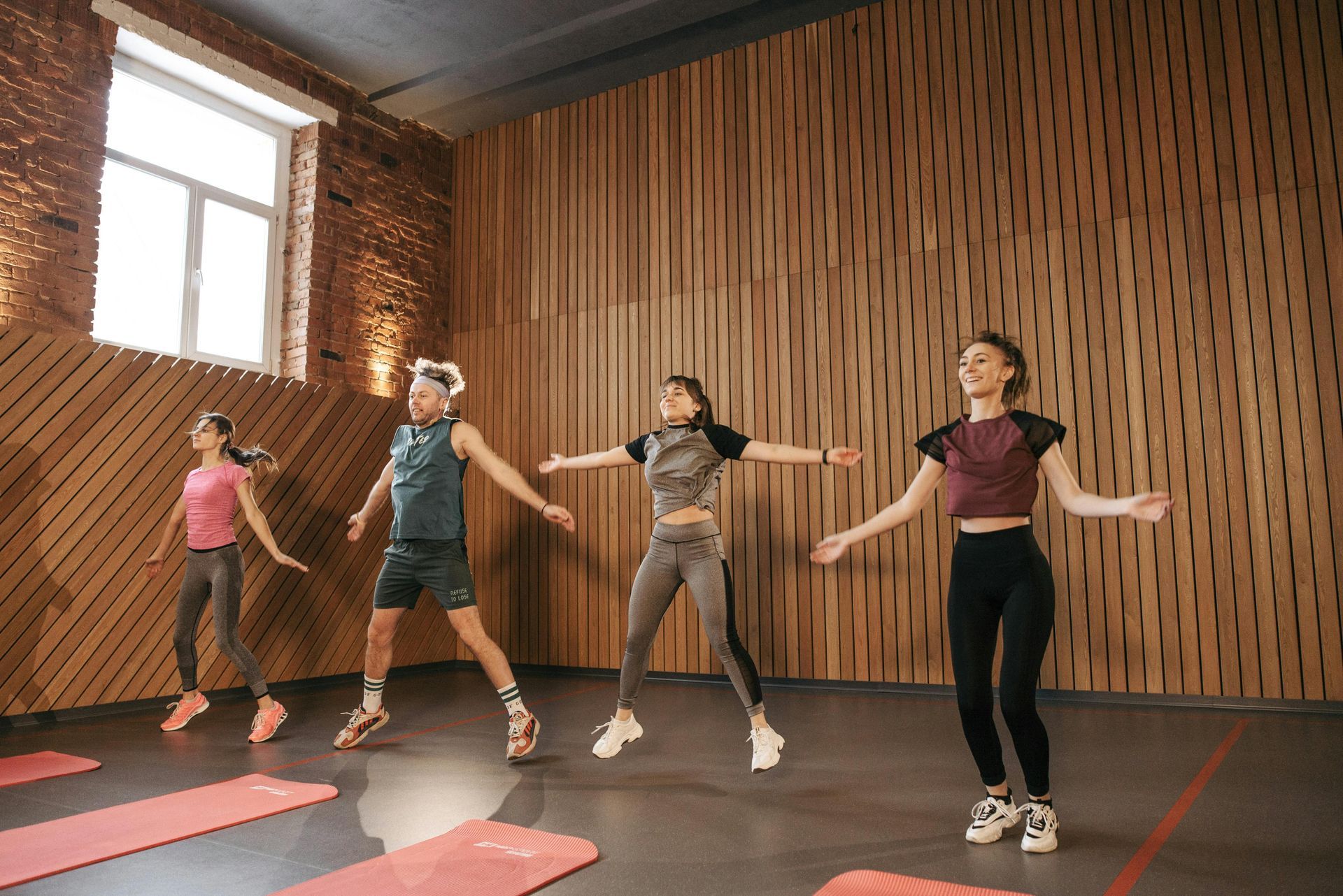 Four people exercising in a dance studio with mats on the floor and a wooden wall backdrop