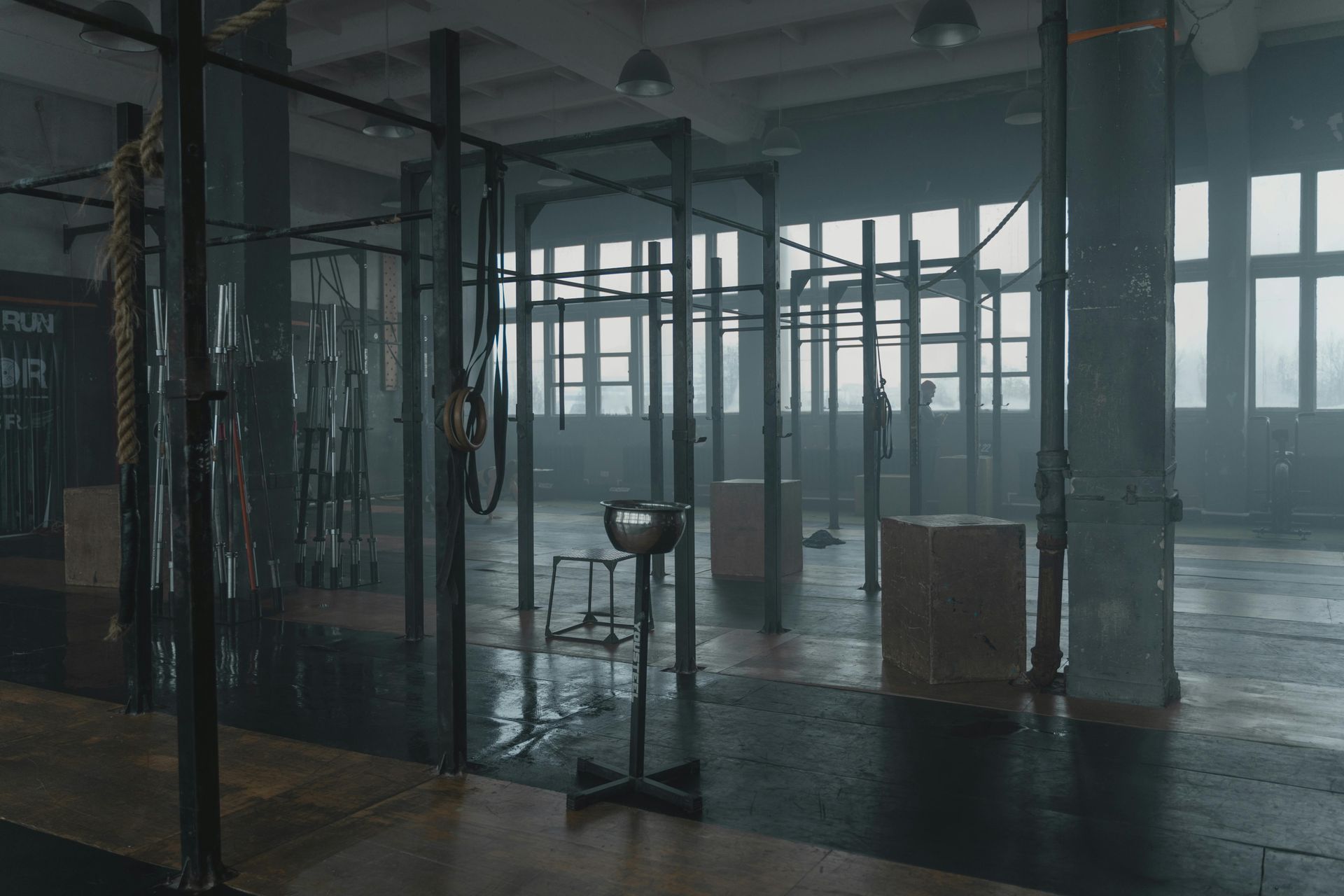 Rain-soaked gym interior with hanging bags, metal frames, and large windows in gray light