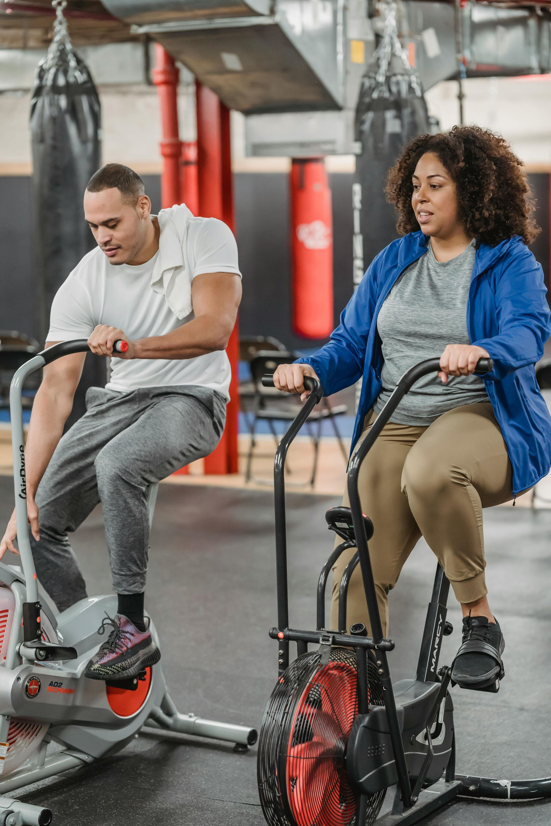 Two people exercising on stationary bikes in a gym, one on a red machine and one on a black machine.