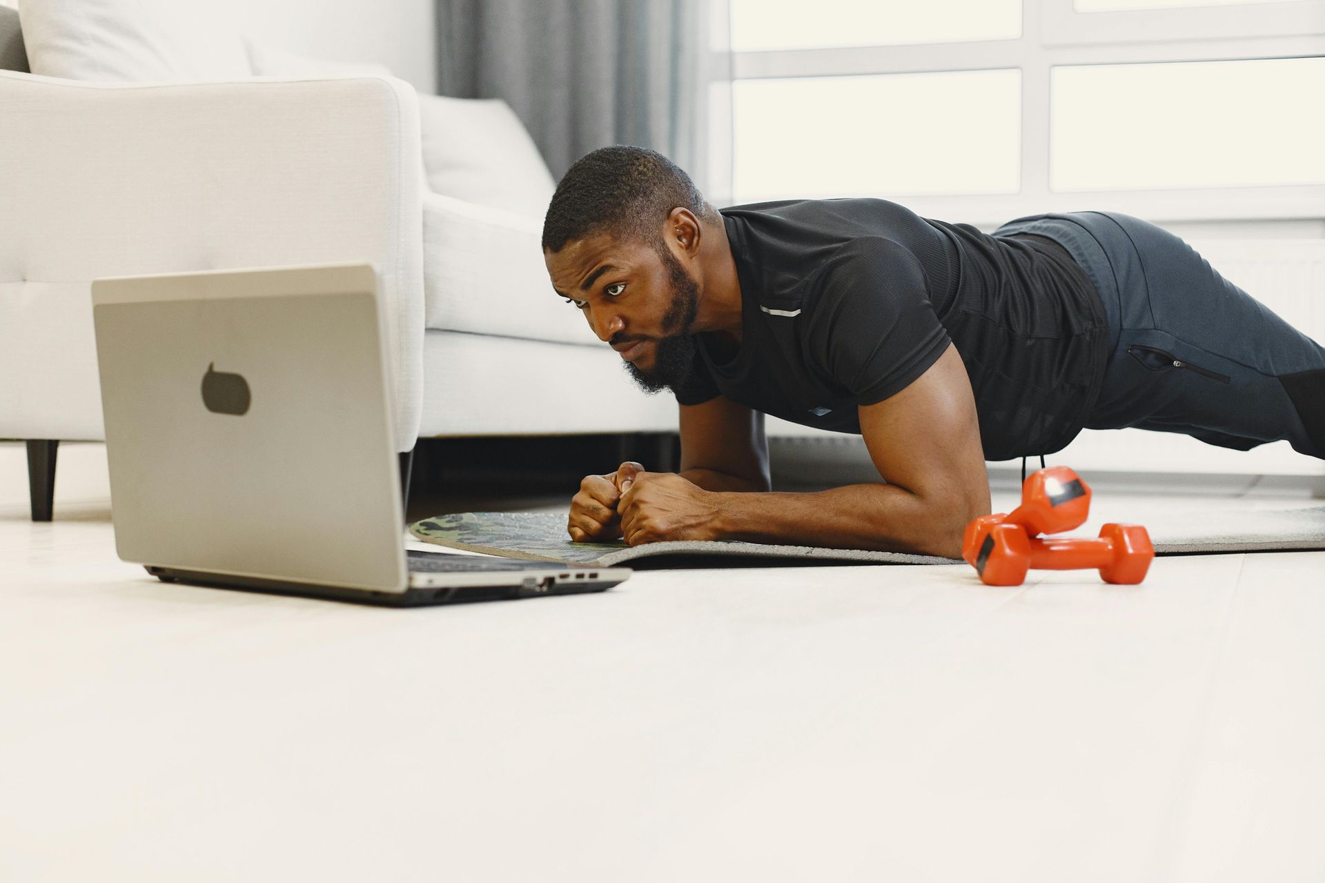Man doing a plank beside a laptop on the floor, with orange dumbbells nearby in a bright room