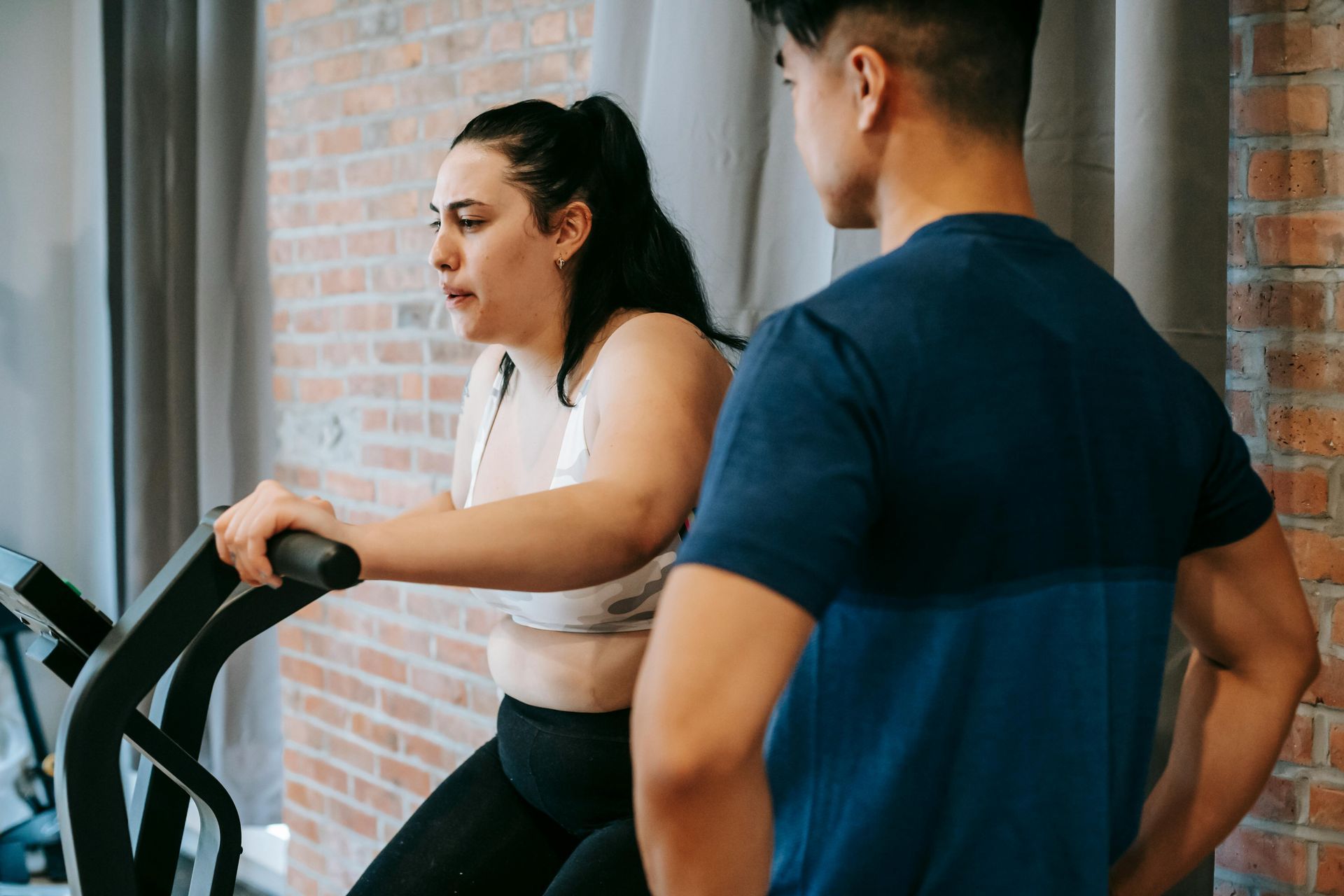 Two people at a gym, one exercising on a machine while the other watches nearby