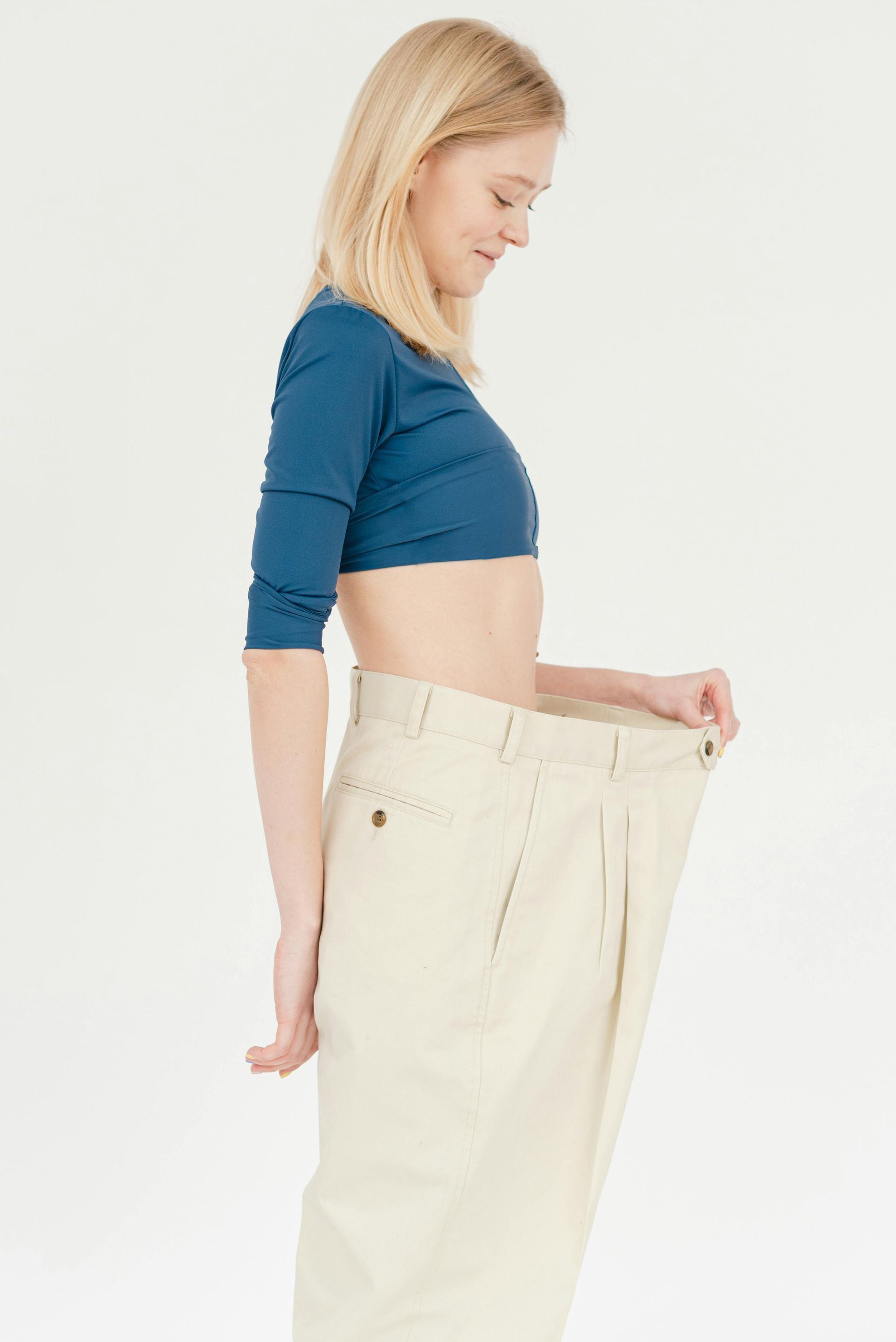 Woman holding out the waistband of oversized cream pants while standing in profile on a white background