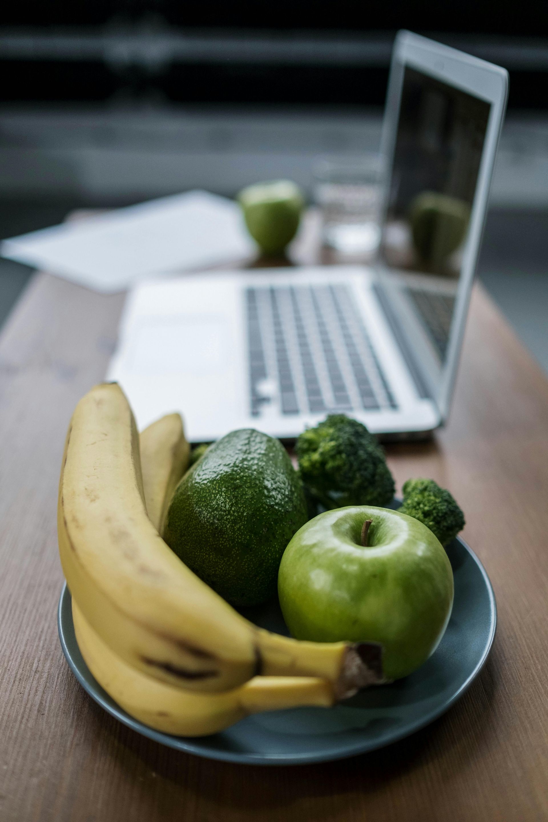 Bananas, avocado, broccoli, and green apple on a plate beside a laptop on a table