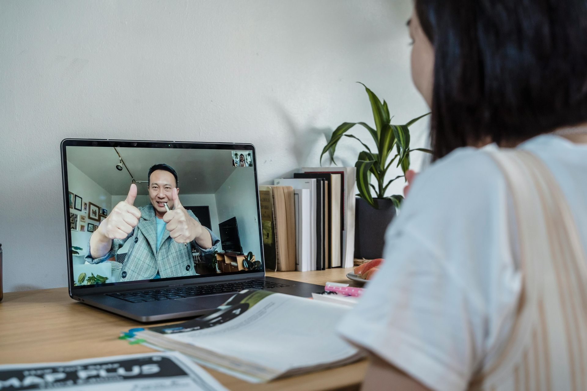 Person on a video call on a laptop, smiling and giving a thumbs-up at a desk with papers and plants