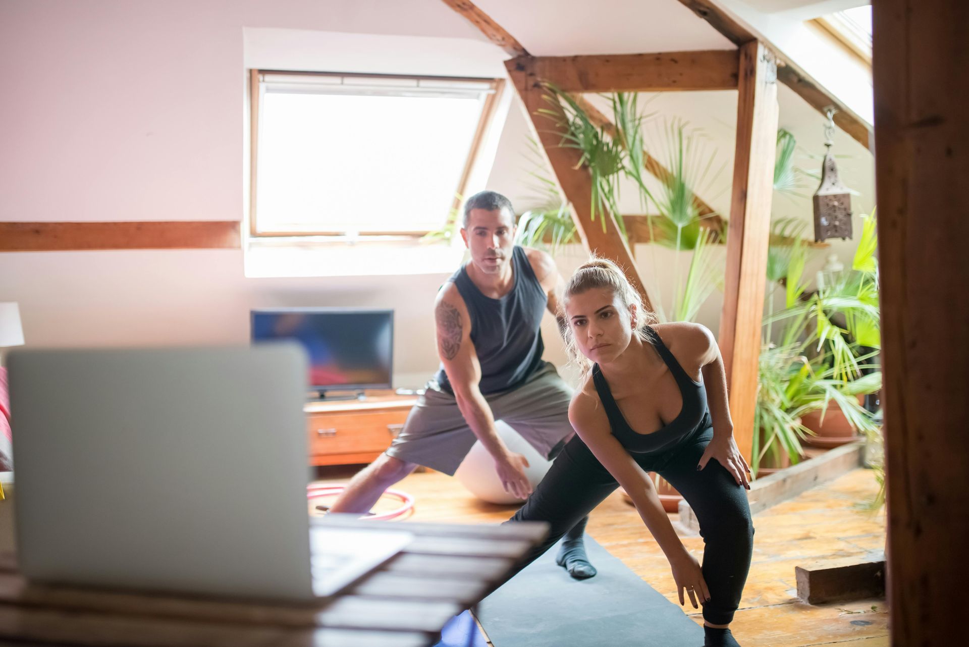 Two people stretching in a bright yoga studio beside a laptop and potted plants