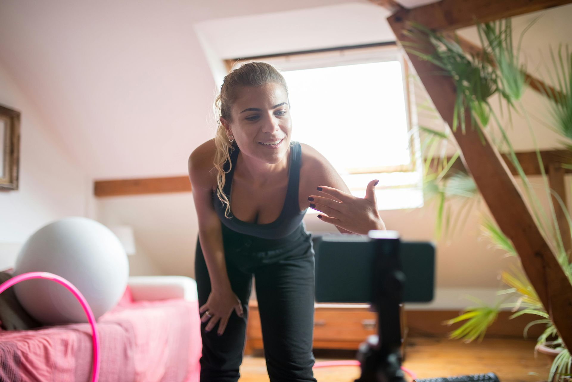 Woman filming a fitness pose in a bright room, leaning toward a phone on a tripod.