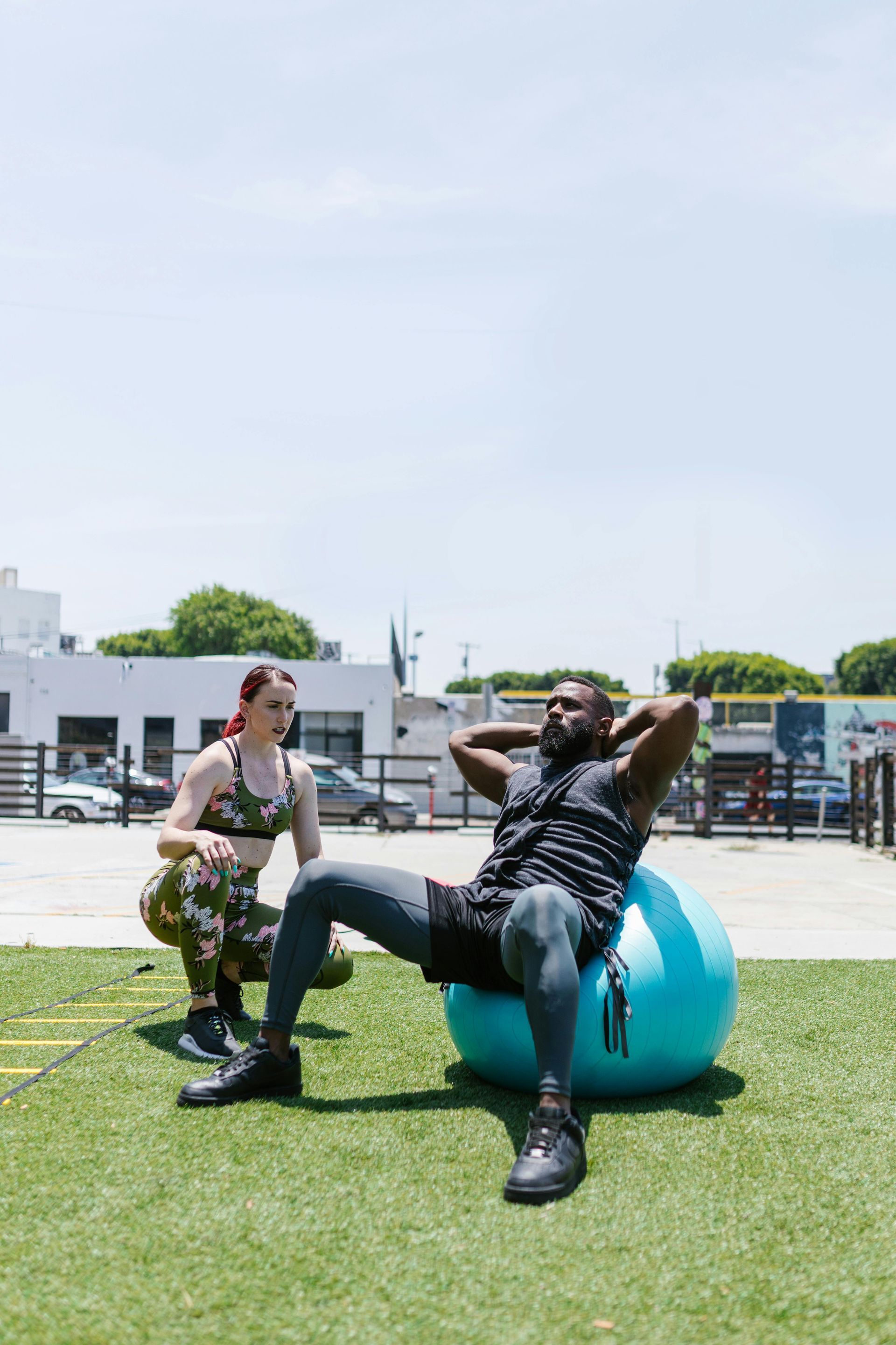 Two people sitting outdoors on a blue exercise ball, one stretching on a grassy lawn.