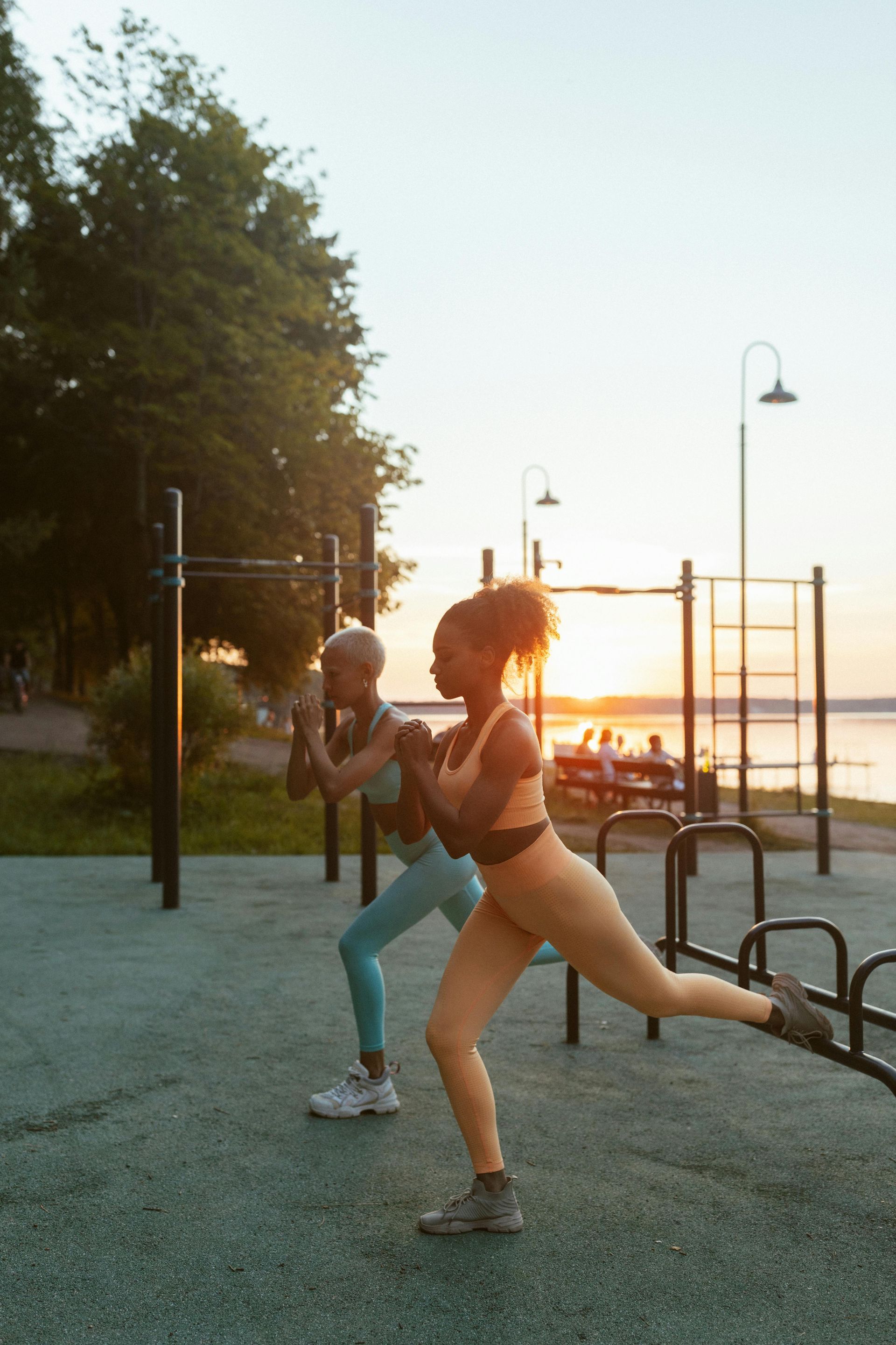 Two people exercise on a waterfront promenade at sunset, one kicking a leg back.