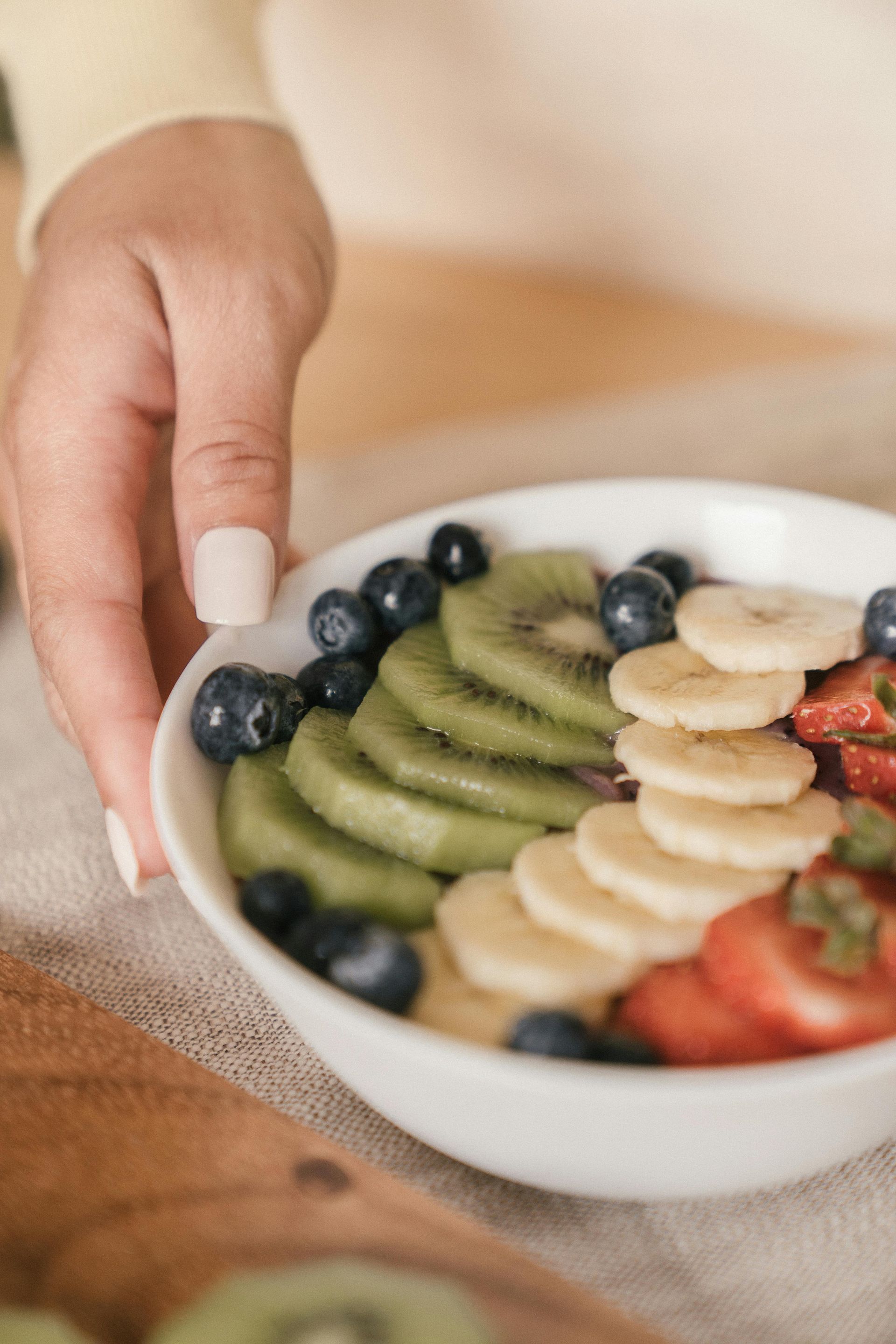 Hand holding a bowl of fruit topped with kiwi, banana, blueberries, strawberries, and oatmeal on a table