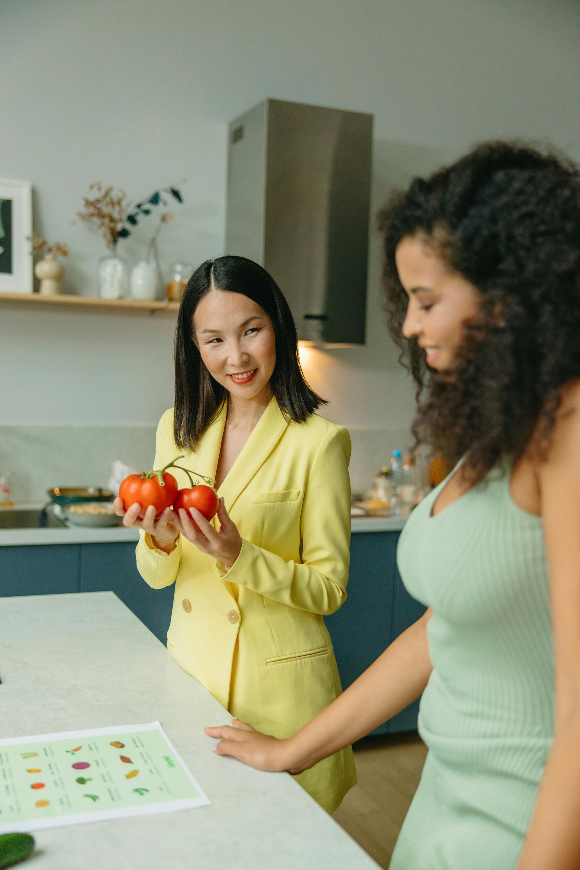 Two women in a kitchen holding tomatoes and looking at a recipe sheet