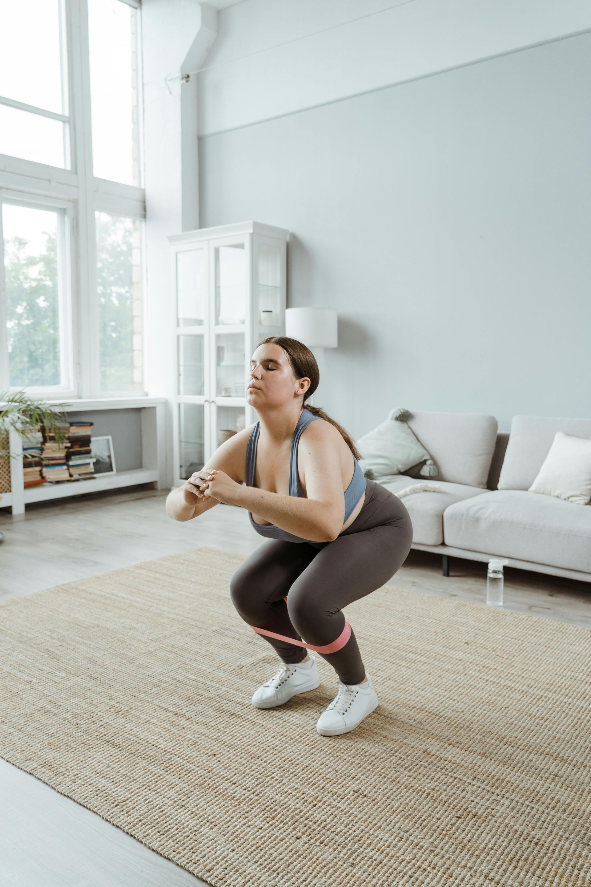 Woman doing a squat workout in a bright living room with a sofa and large windows.