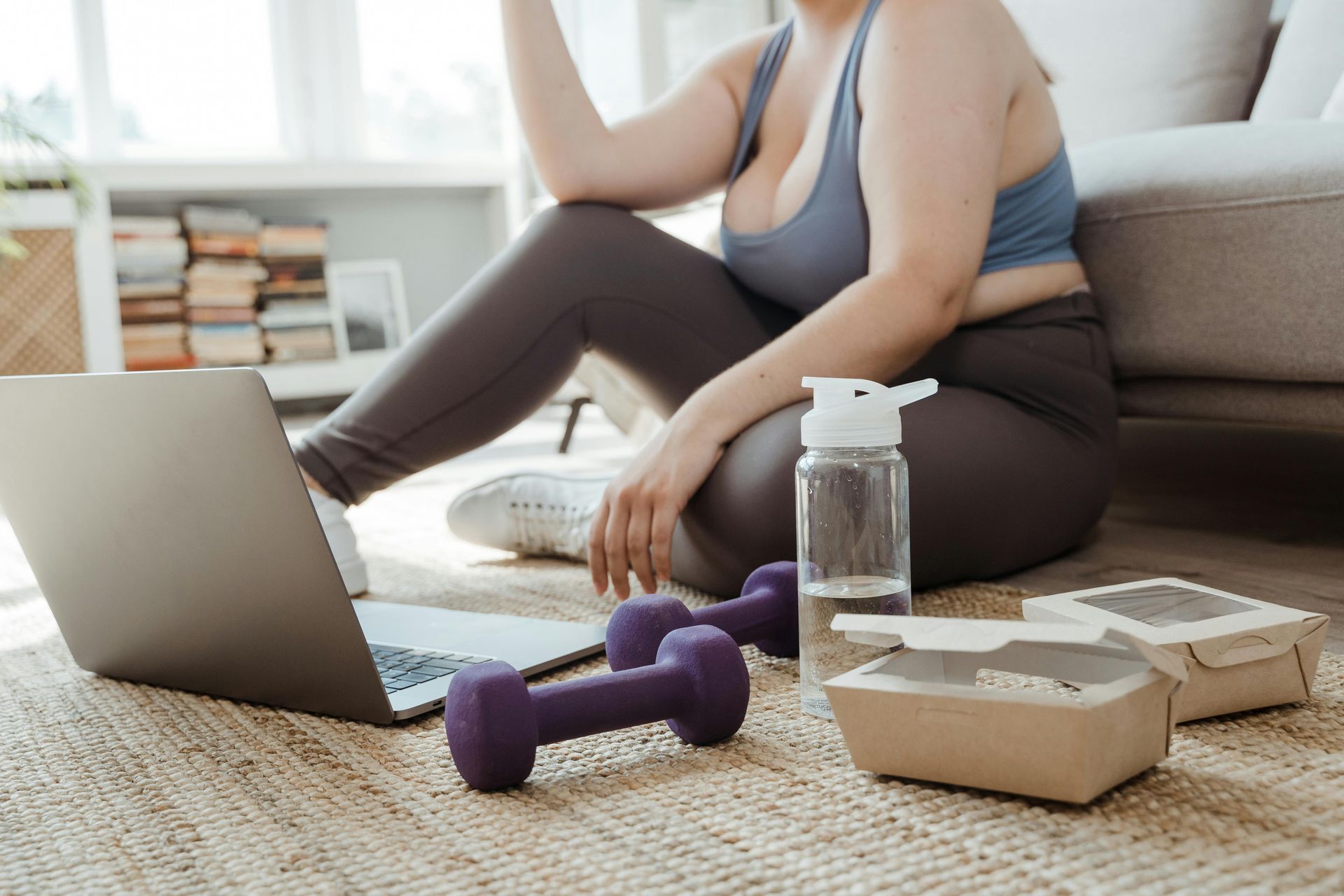 Person stretching beside a laptop, water bottle, dumbbell, and takeout box on a carpeted floor