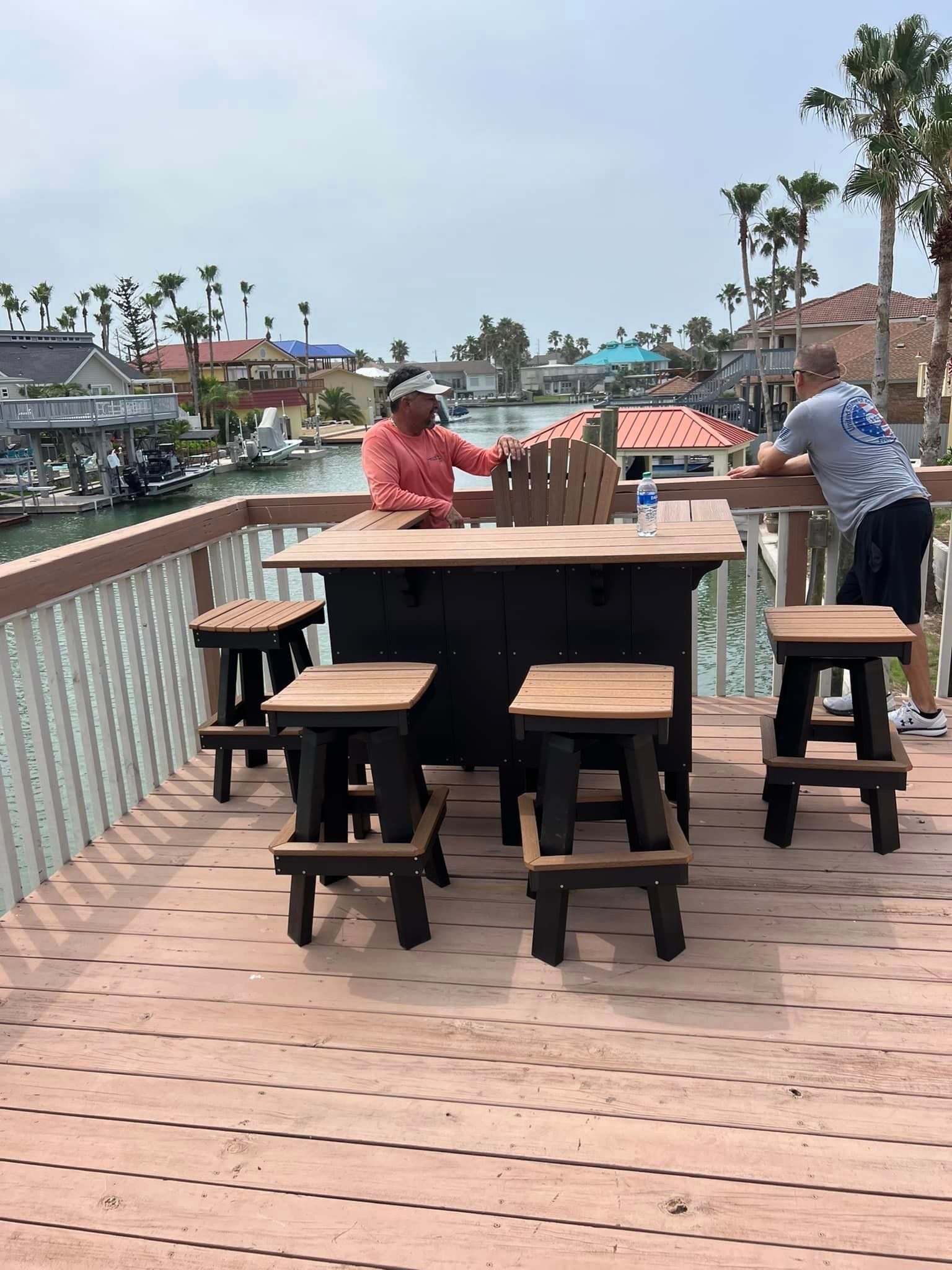 A man is sitting at a table with stools on a deck overlooking a body of water.