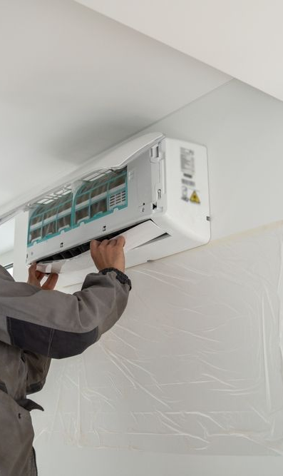 Person opening an air conditioning unit for maintenance, indoors. Unit is mounted on a white wall with plastic protective sheeting.