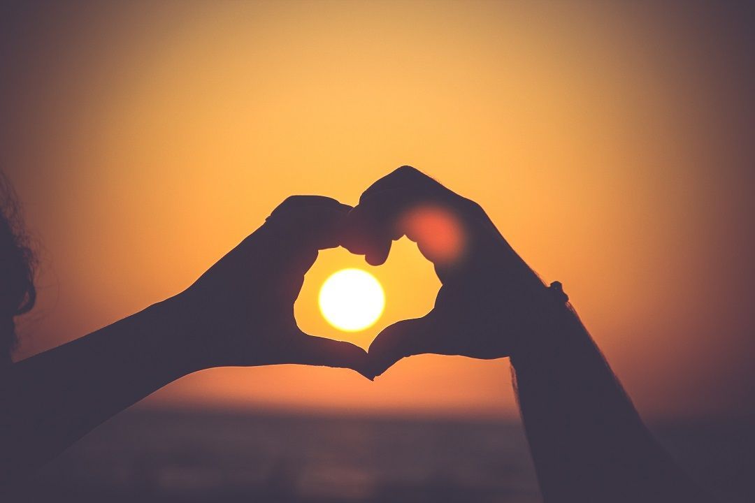 a person is making a heart shape with their hands in front of the sun at sunset .
