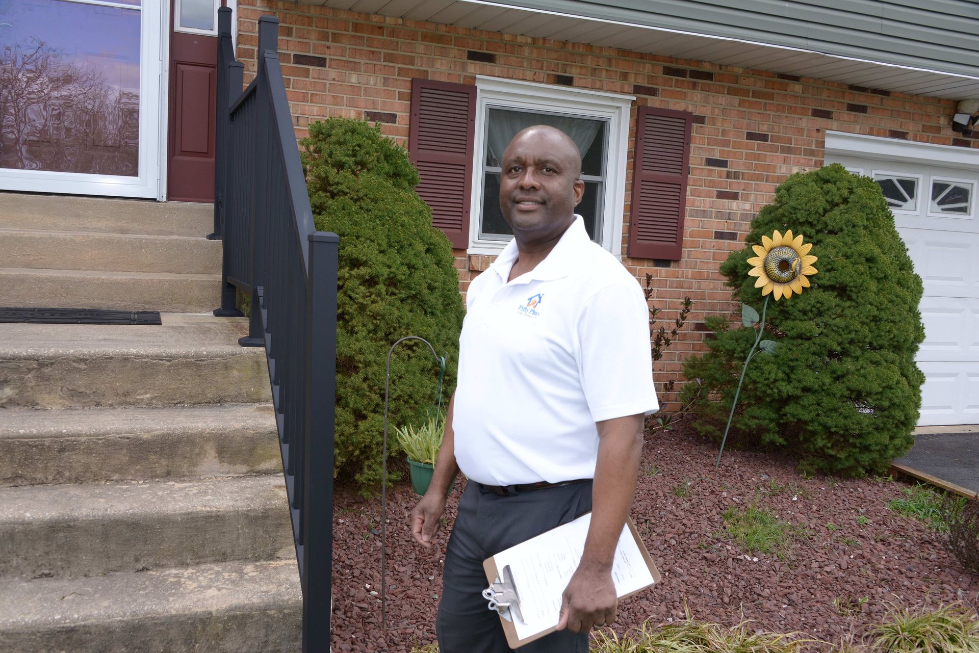 A person in a white polo shirt holds a clipboard while standing on the steps of a brick house with a sunflower decoration.
