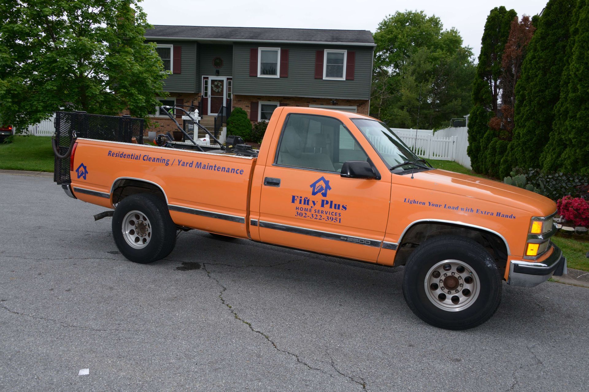 An orange pickup truck with company branding parked on an asphalt street in front of a two-story suburban house.