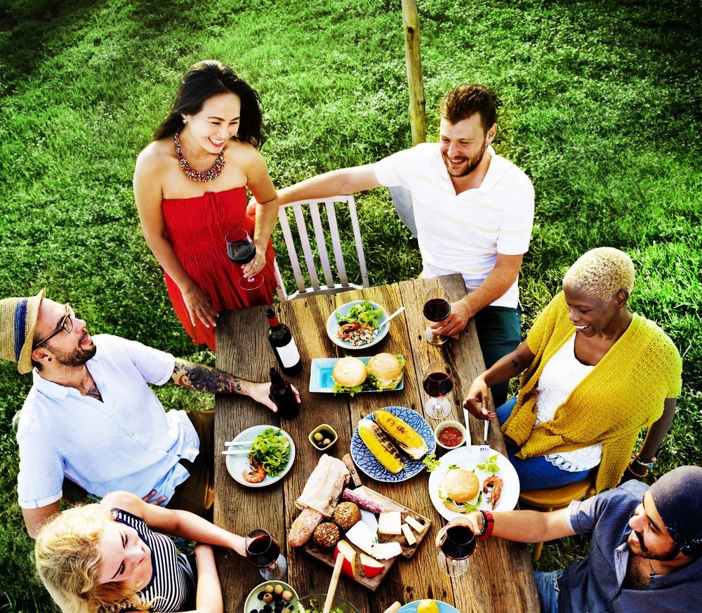 Five people sit around a wooden outdoor table with food and drinks, laughing and talking in a grassy setting.