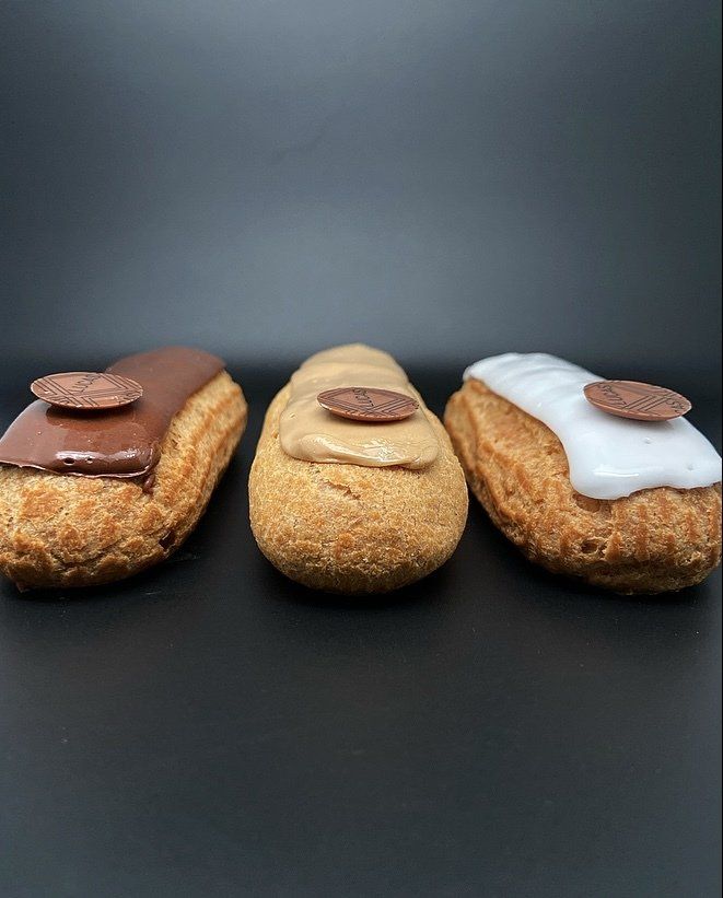 Trois éclairs avec un glaçage de couleurs différentes, chacun surmonté d'un disque de chocolat, sur une surface noire.
