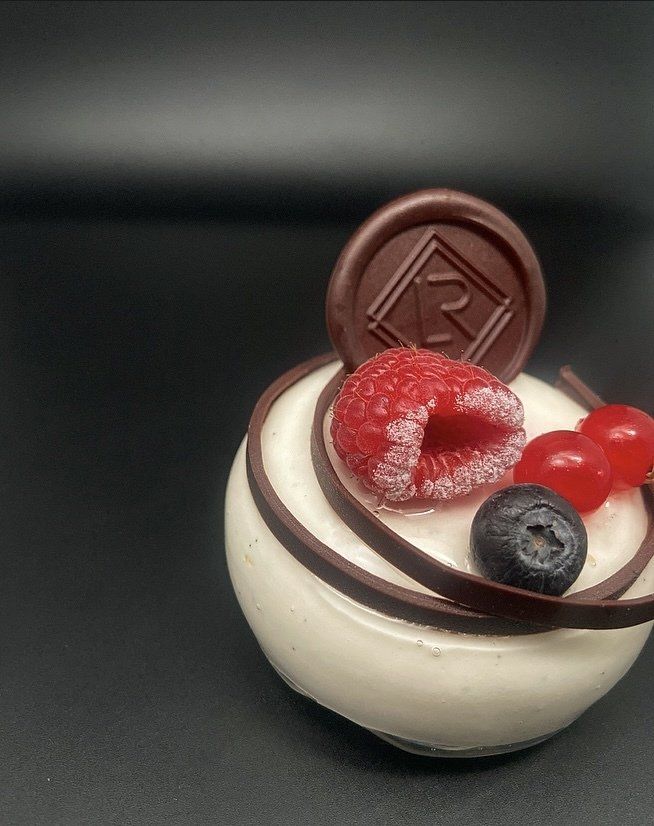 Un dessert blanc et rond avec un tourbillon de chocolat, surmonté d'un sceau de framboise, de myrtille et de chocolat sur un fond noir.