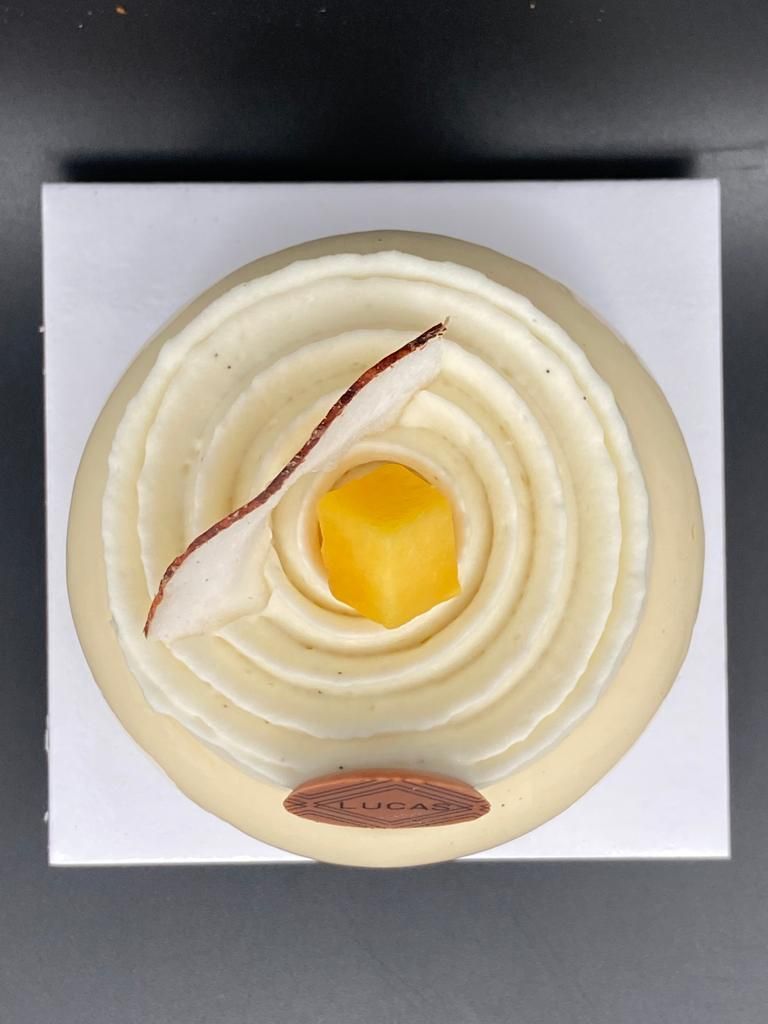 Gâteau glacé blanc avec un motif en spirale, surmonté d'un cube jaune et de flocons de noix de coco, sur une boîte carrée blanche.
