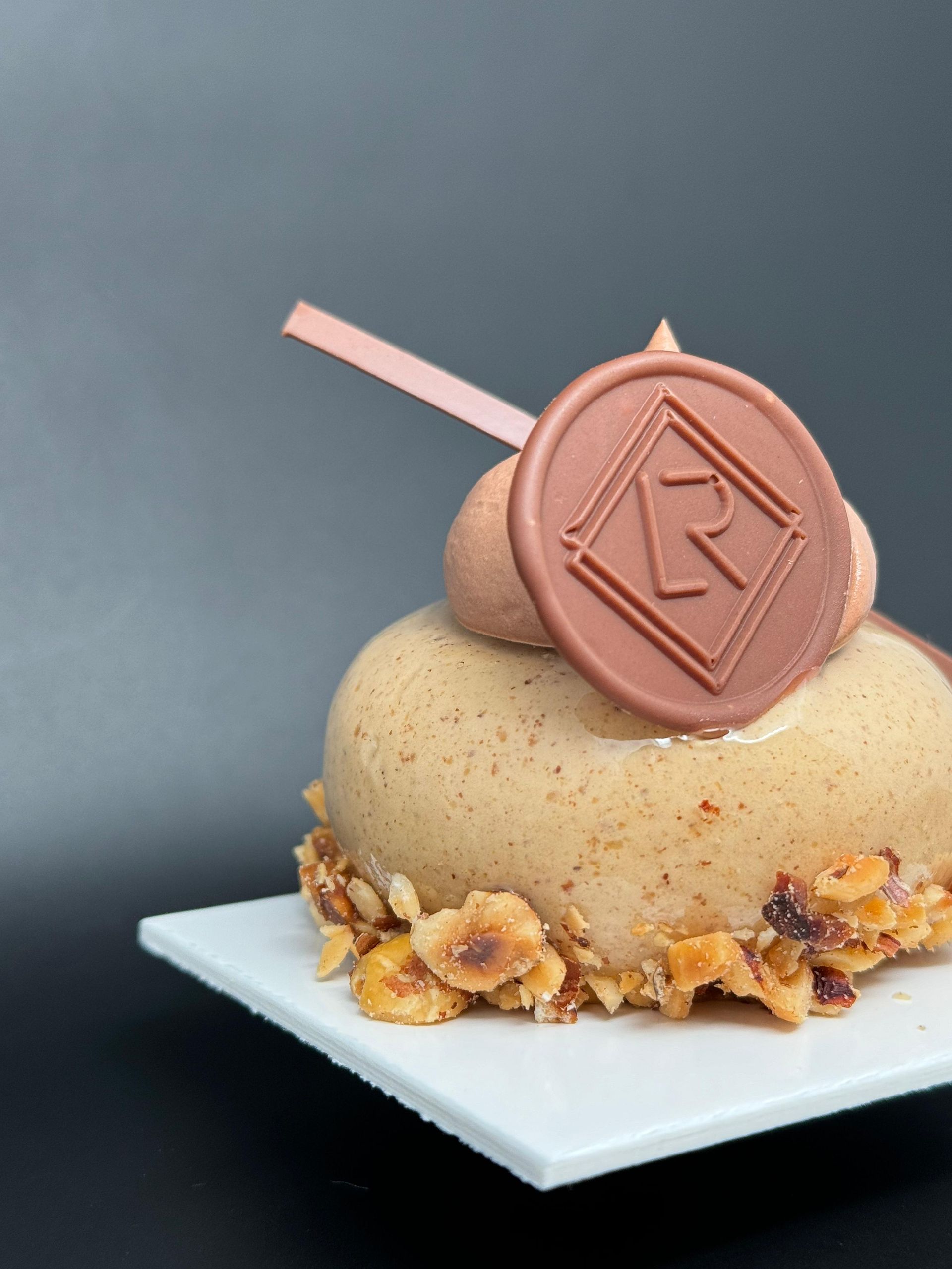 Dessert gourmand sur assiette carrée blanche; croûte de noisettes, glaçage moka, médaillon logo et garniture rose.