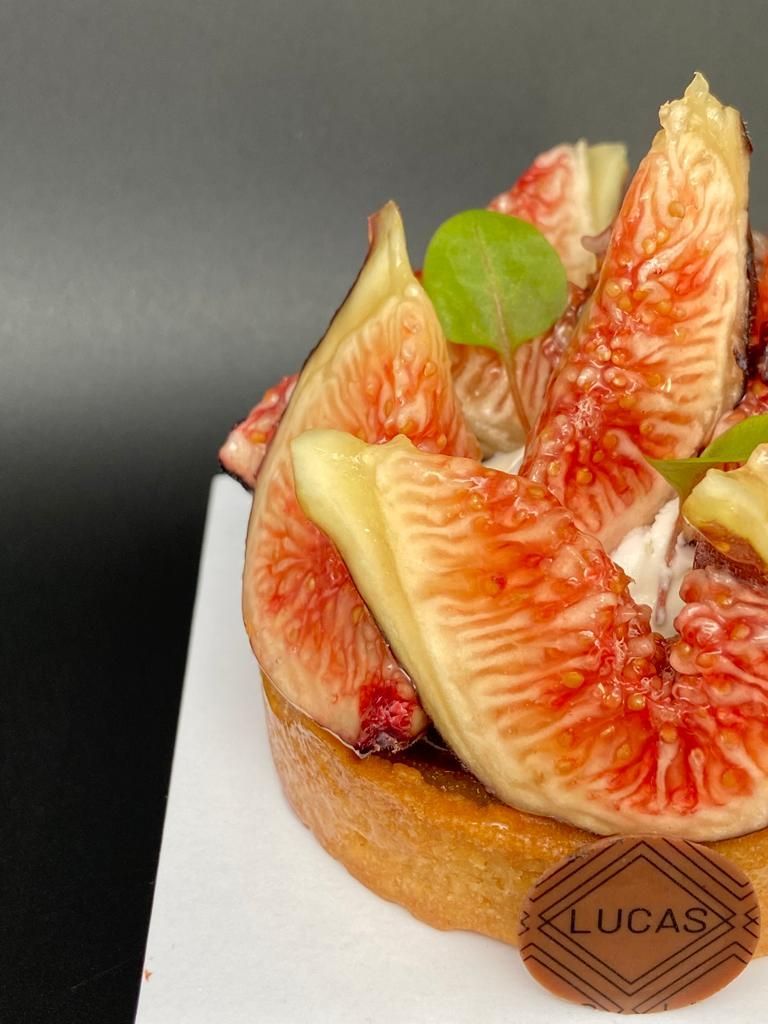 Gros plan d'une tarte aux figues avec des figues tranchées, de la crème et un logo LUCAS sur une base blanche sur fond noir.