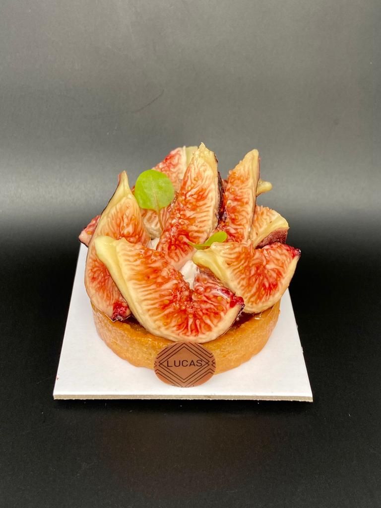 Tarte aux figues sur une assiette carrée blanche, garnie de figues tranchées, d'une feuille et d'un logo en chocolat, sur fond noir.