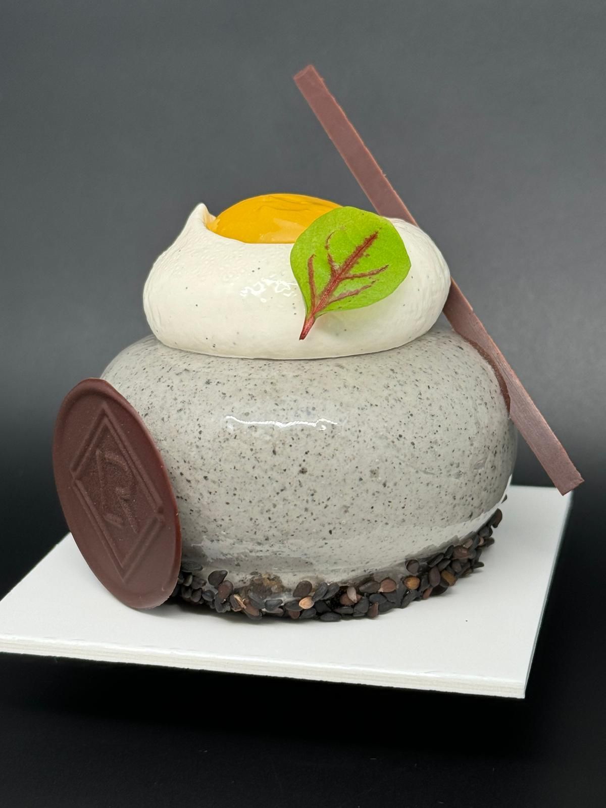 Une pâtisserie gourmande avec un glaçage gris, garnie de crème blanche, de fruits et d'un sceau en chocolat sur une assiette blanche.